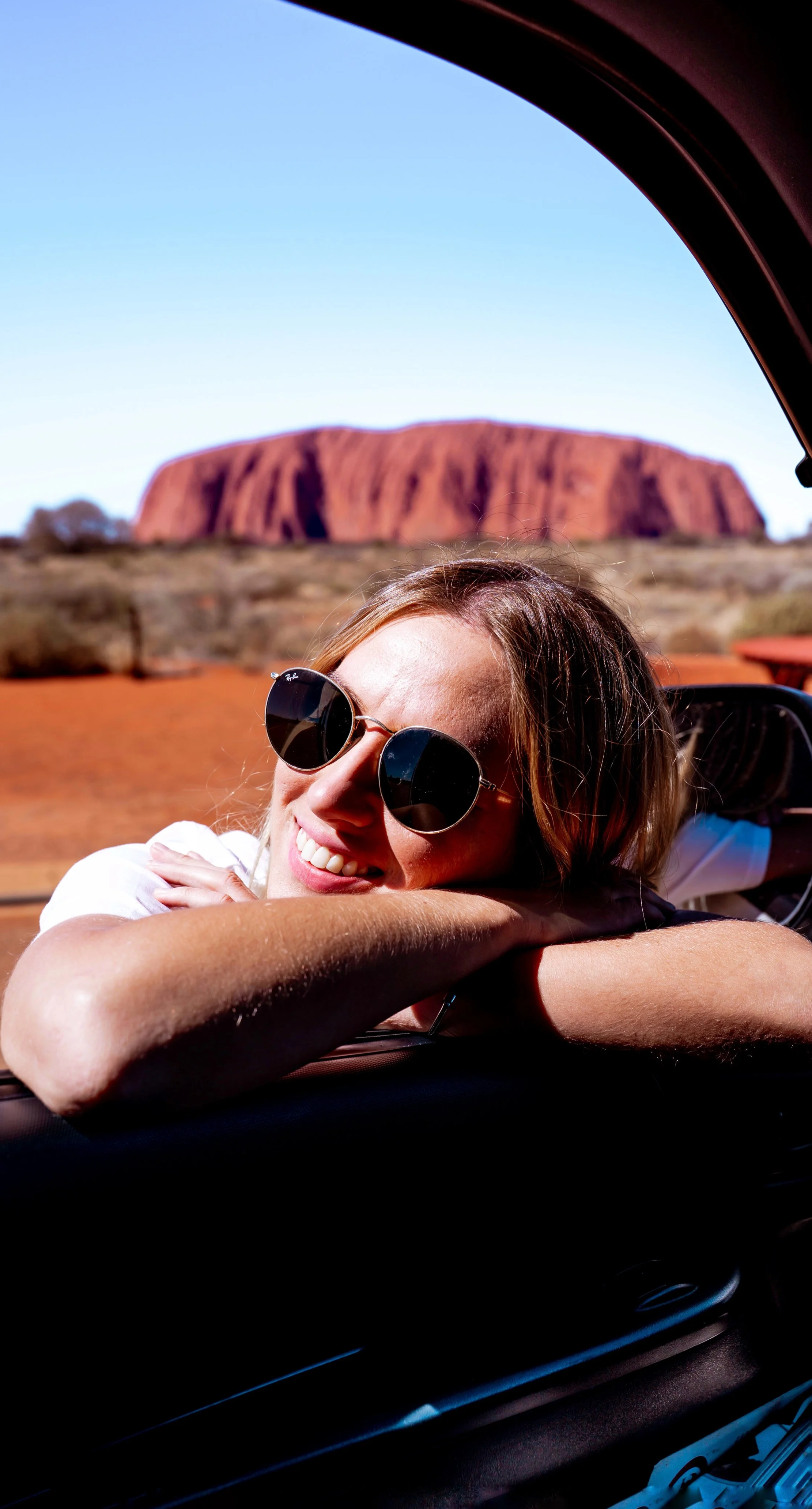 A woman with sunglasses smiling and resting her head on the window ledge of a car, with Uluru in the background under a clear blue sky.