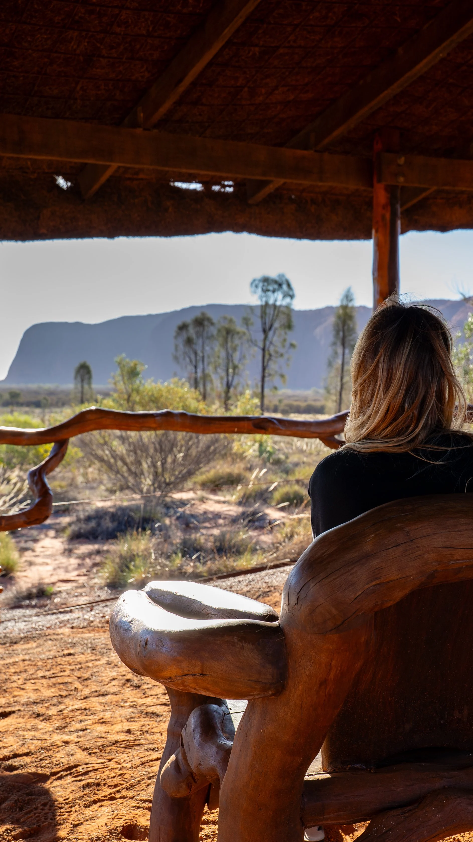 A woman sitting on a wooden bench on a porch, overlooking a desert landscape with bushes, trees, and mountains in the background.