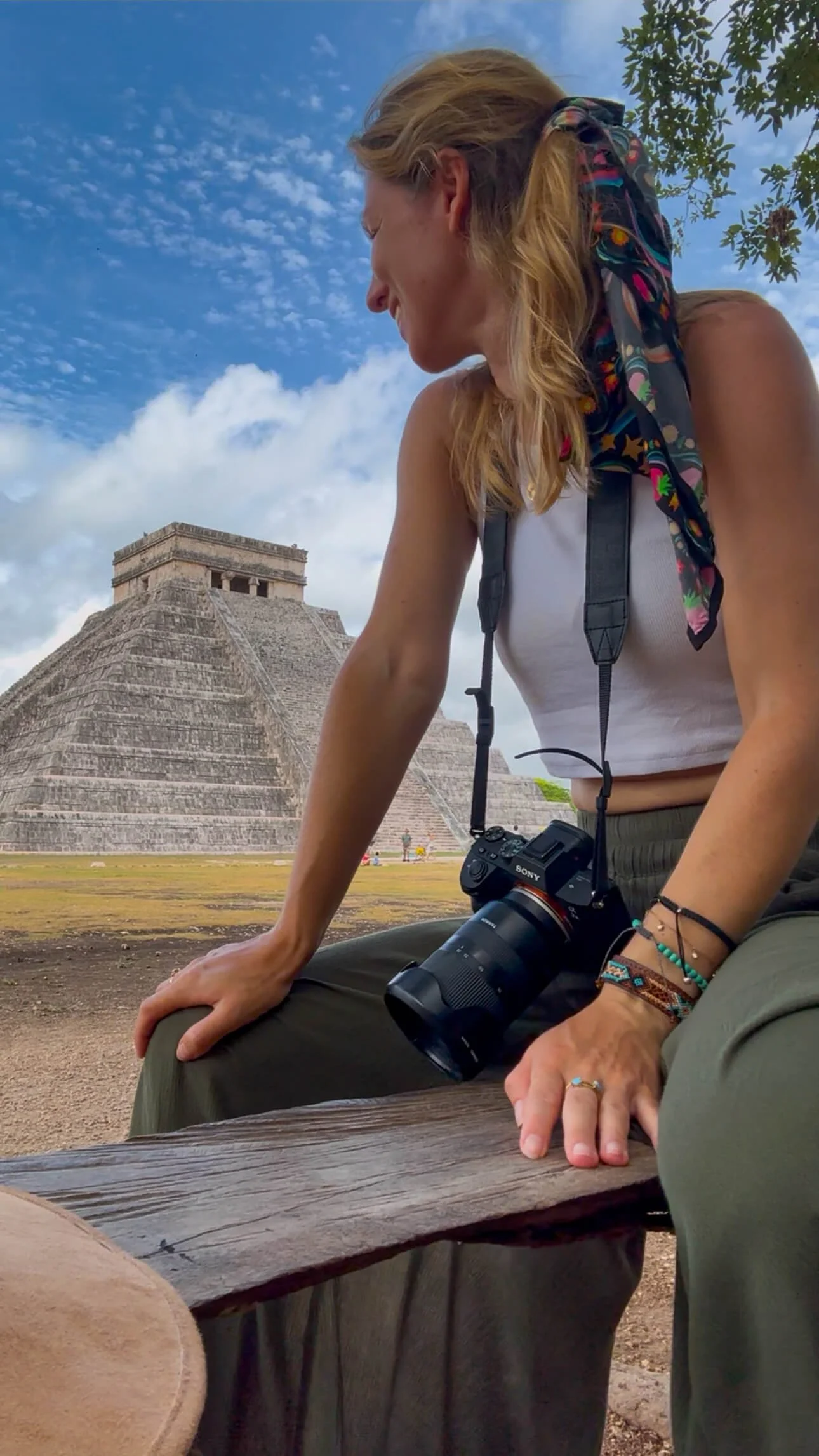 A woman with a camera around her neck, sitting on a wooden bench near Chichen Itza in Mexico, looking at the pyramid structure with a blue sky and some clouds in the background.