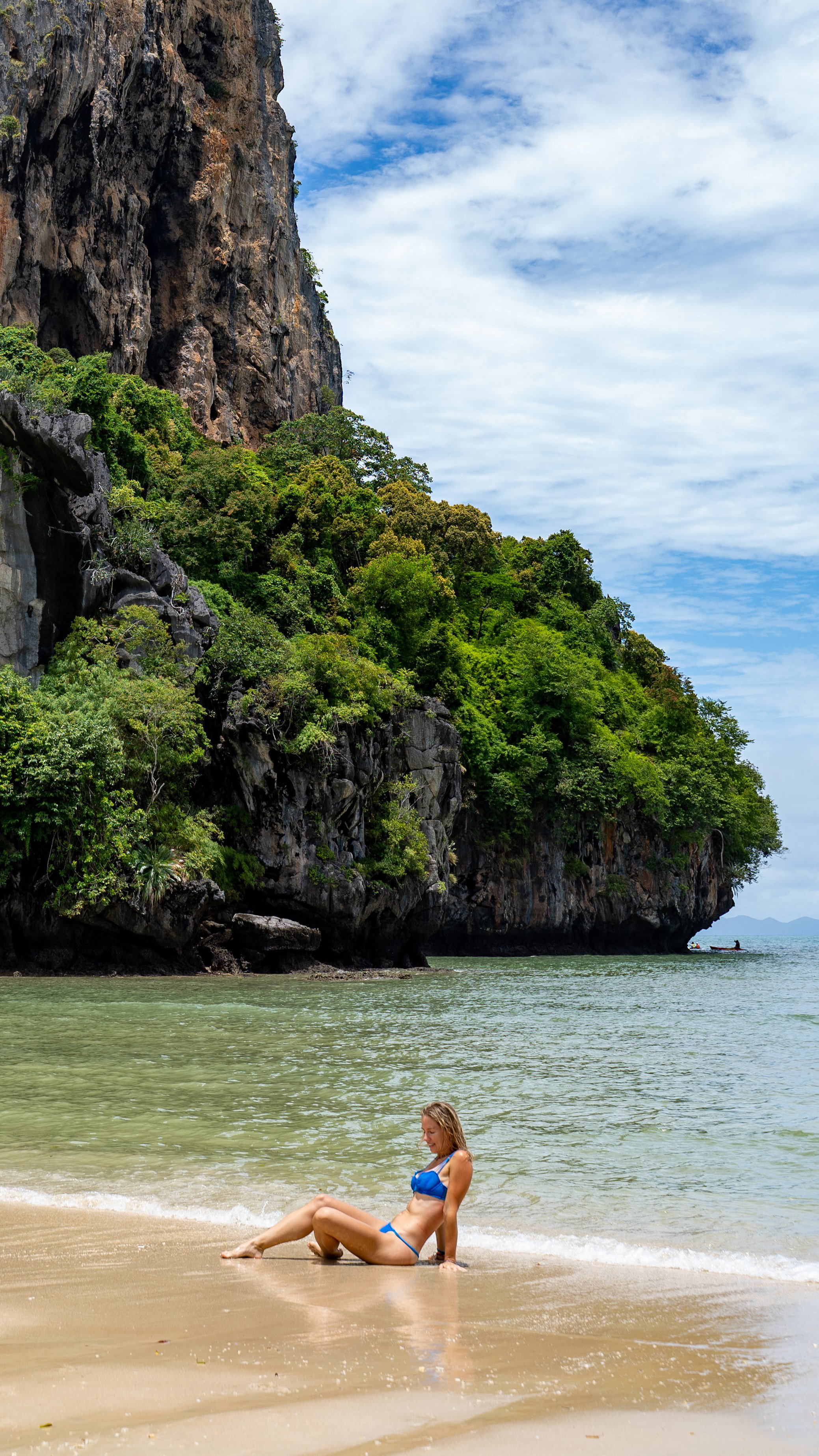 A woman in a blue bikini sitting on the sandy beach near the water, with a large rocky cliff covered in green vegetation and a cloudy blue sky in the background.