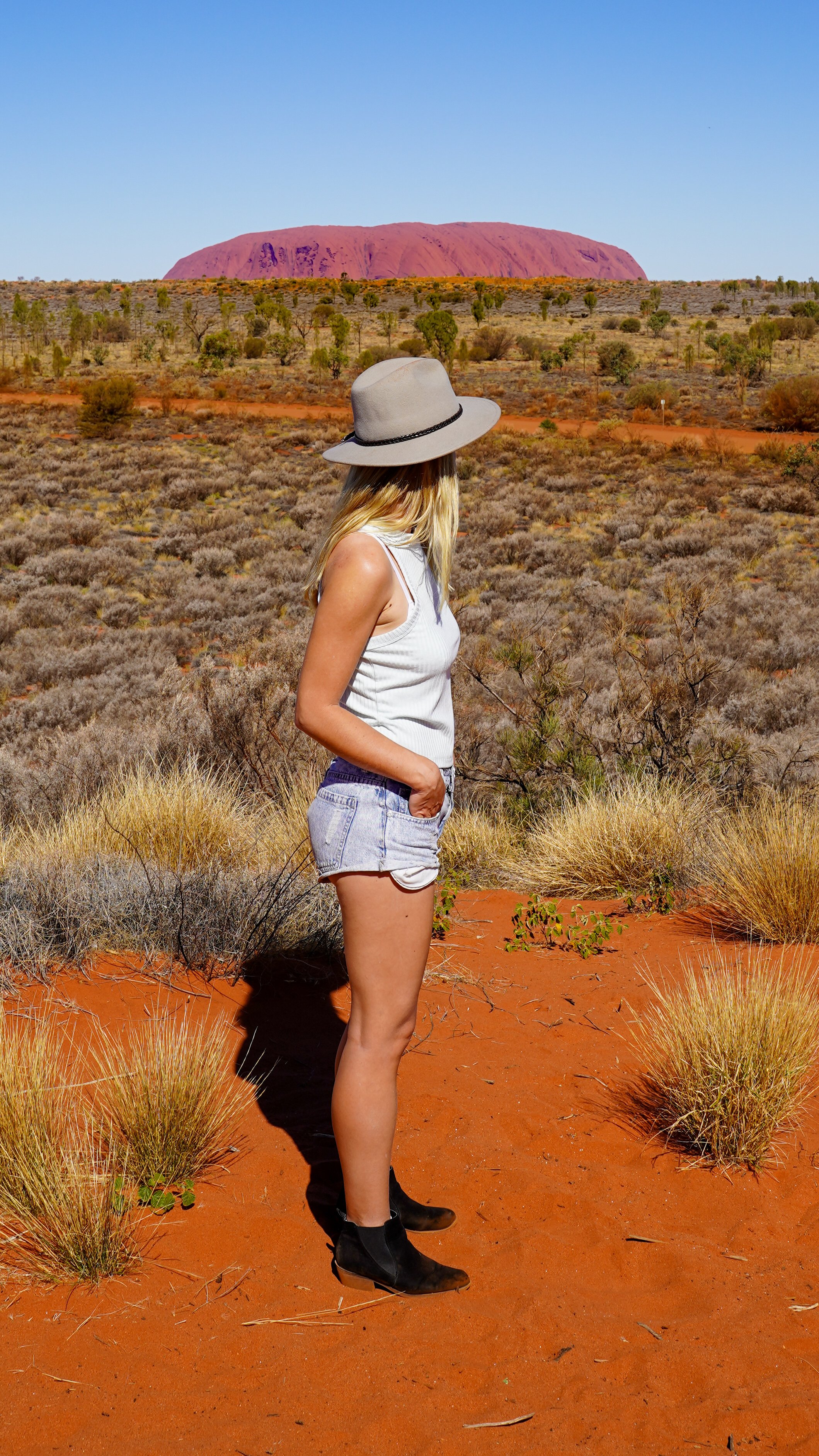 A woman standing in a desert landscape with orange sand, bushes, and sparse vegetation. She is wearing a wide-brimmed hat, sleeveless top, denim shorts, and black ankle boots, looking towards Uluru in the distance.