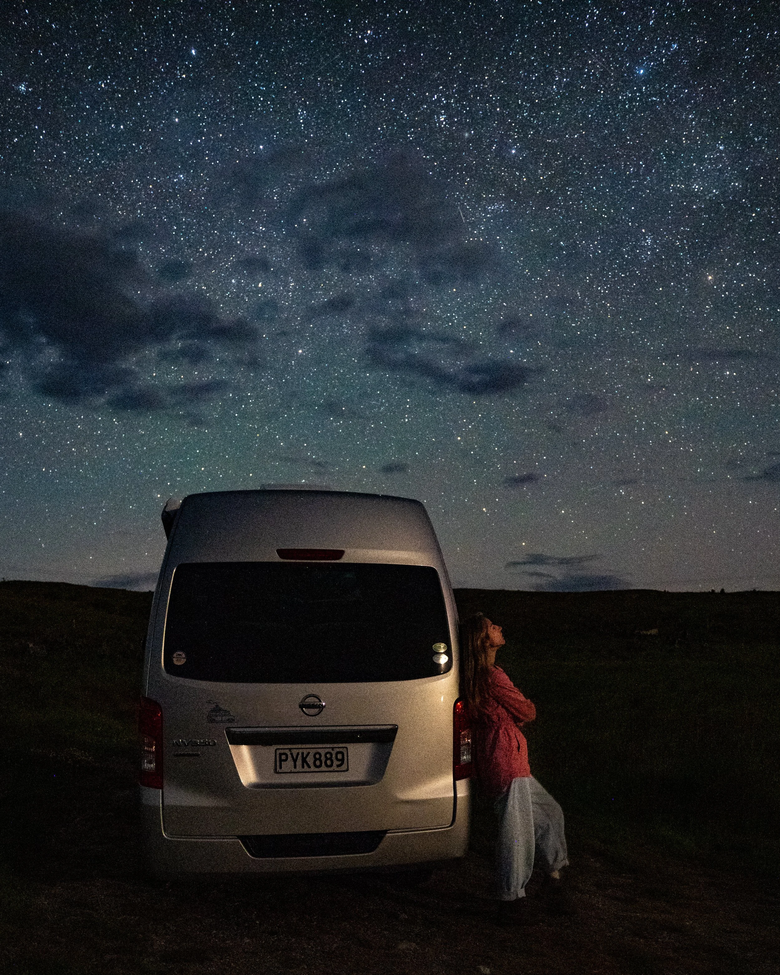 A woman standing next to a white van, looking up at a starry night sky with scattered clouds and a faint shooting star.