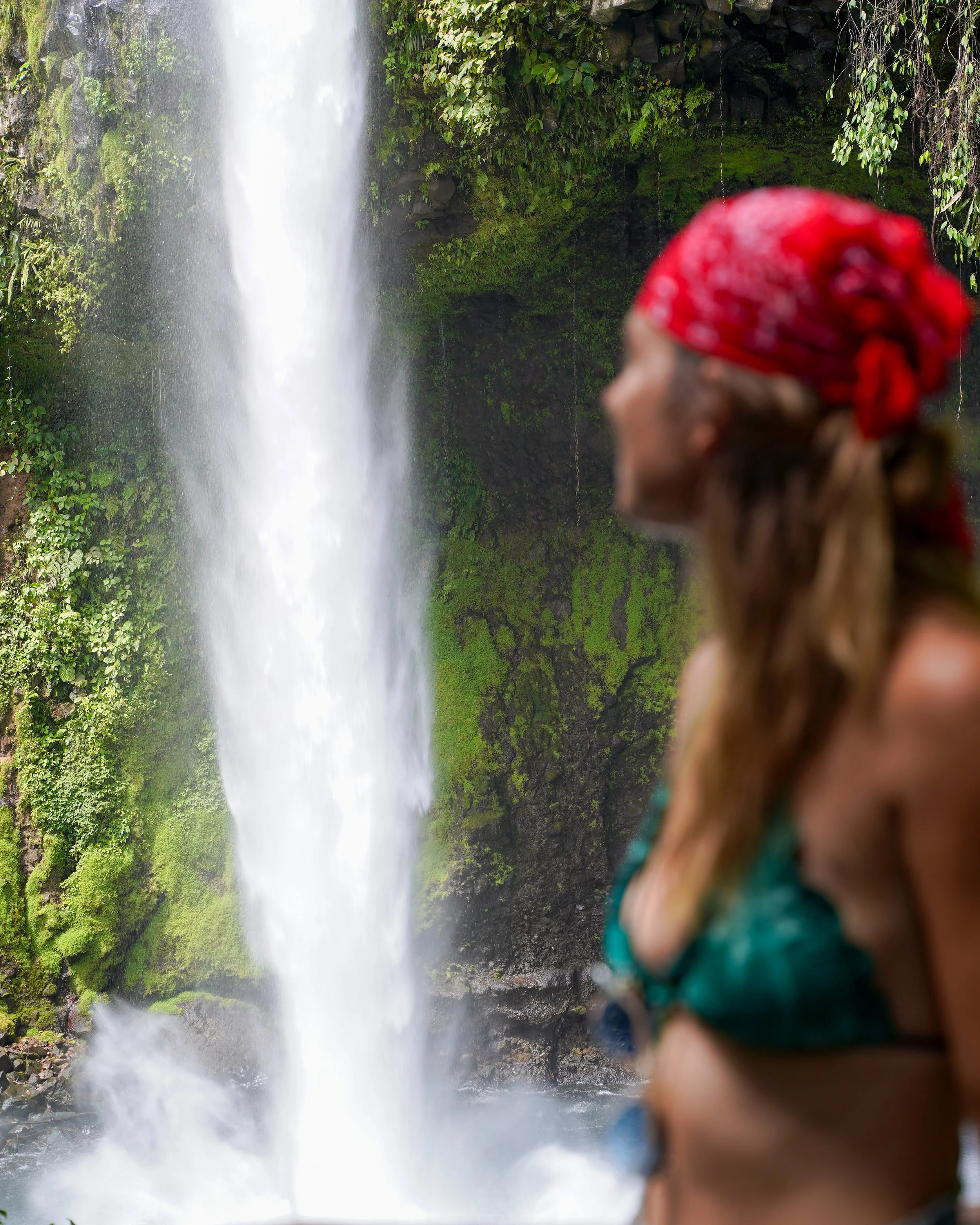 A woman with a red bandana looks at a tall waterfall surrounded by lush green moss and foliage.