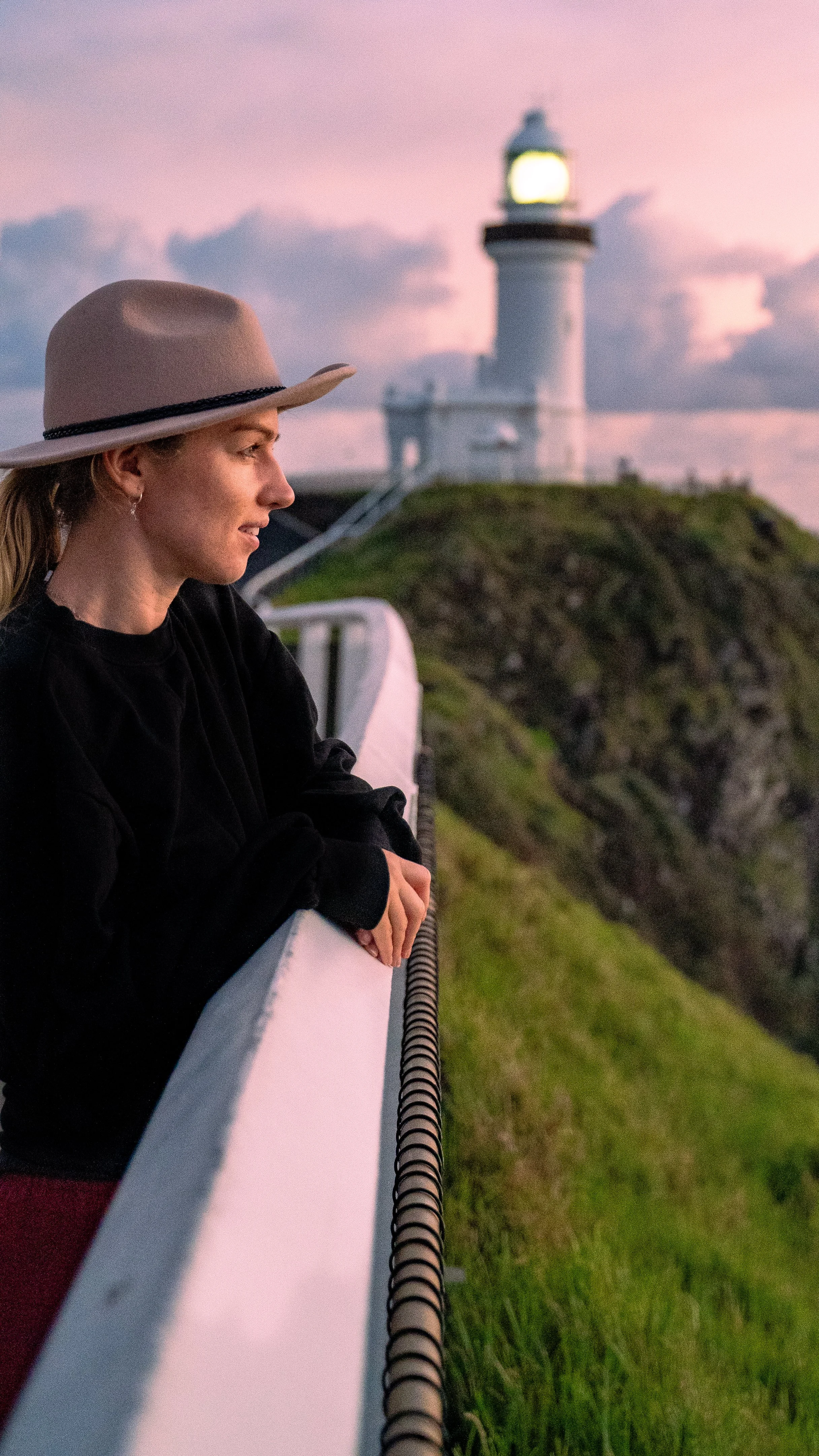 A woman wearing a gray hat and black sweater leaning on a white railing, looking at the lighthouse in the distance during dusk.
