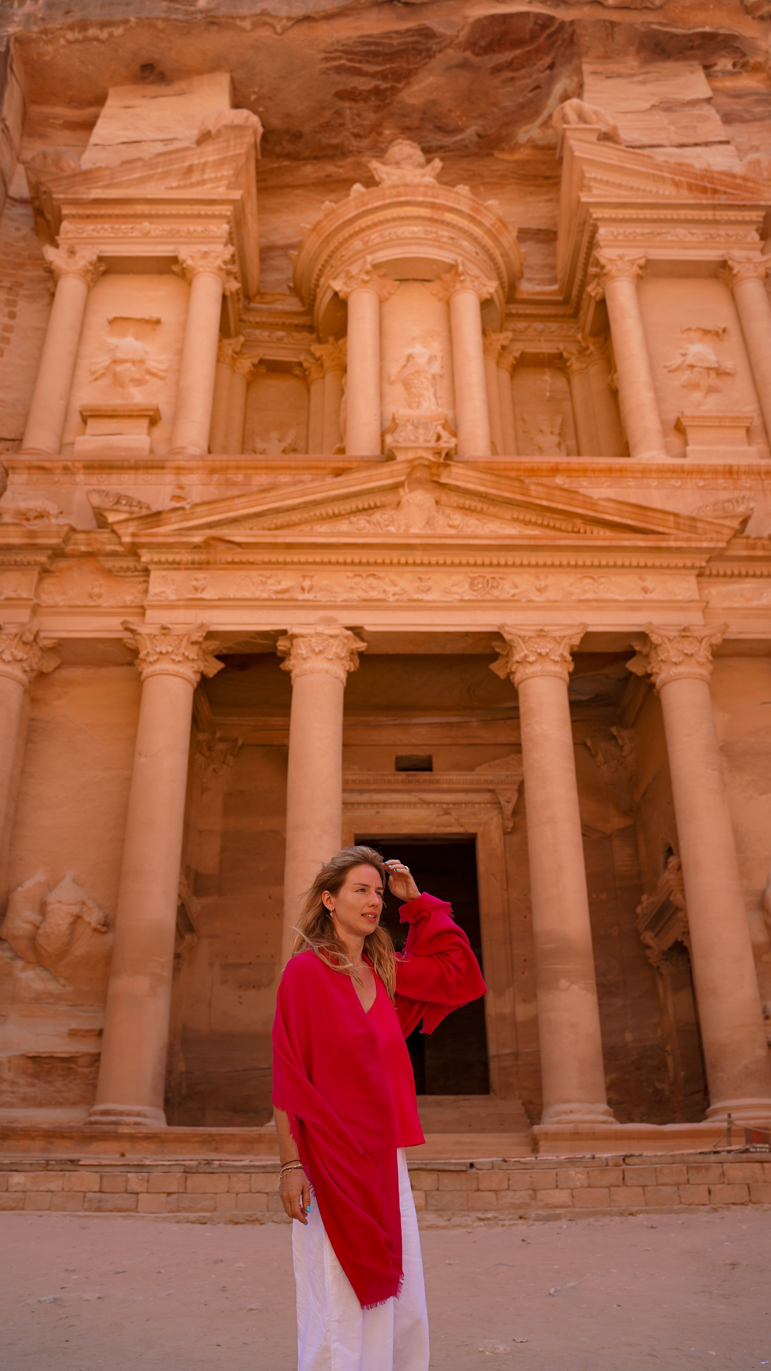 A woman wearing a red shawl and white pants standing in front of the Al-Khazneh (Treasury) monument in Petra, Jordan.