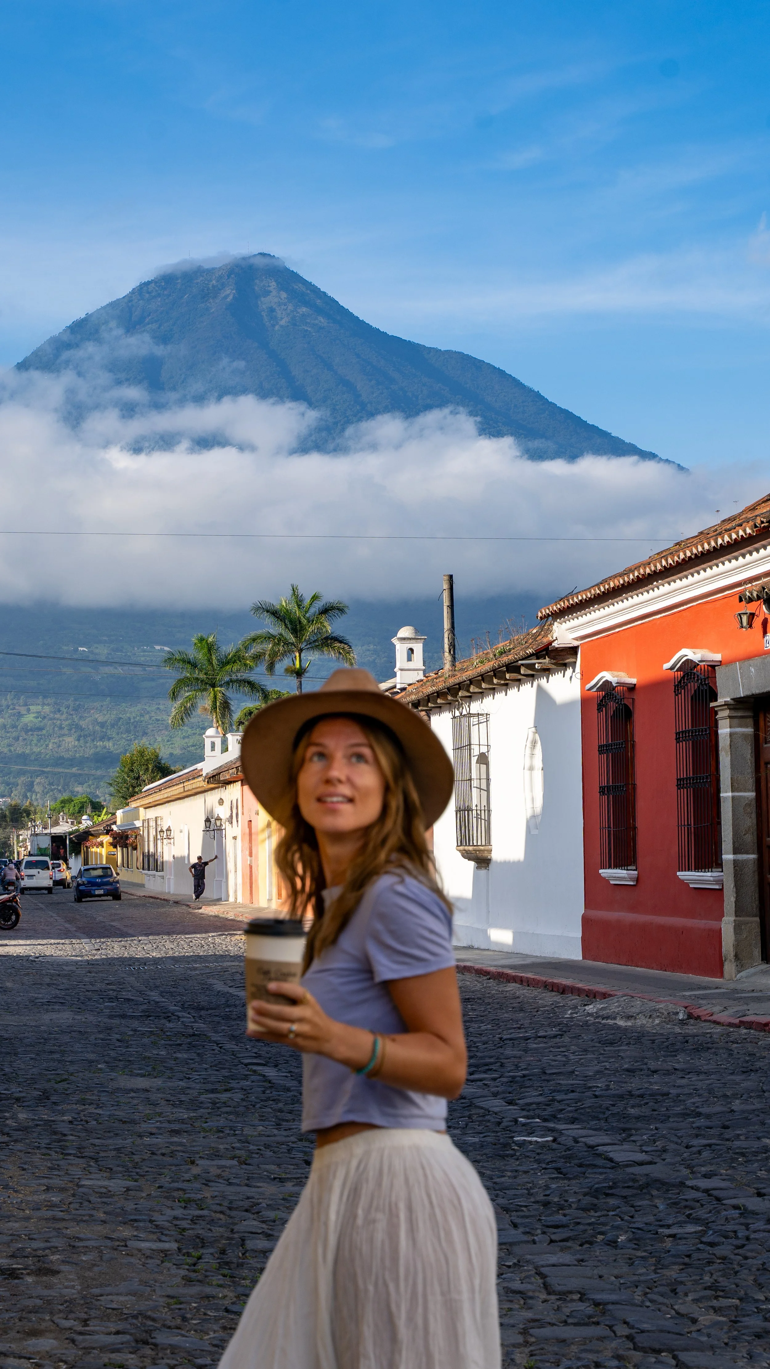 A woman wearing a wide-brimmed hat holding a coffee cup standing on a cobblestone street with colonial-style buildings, palm trees, and a volcano in the background.