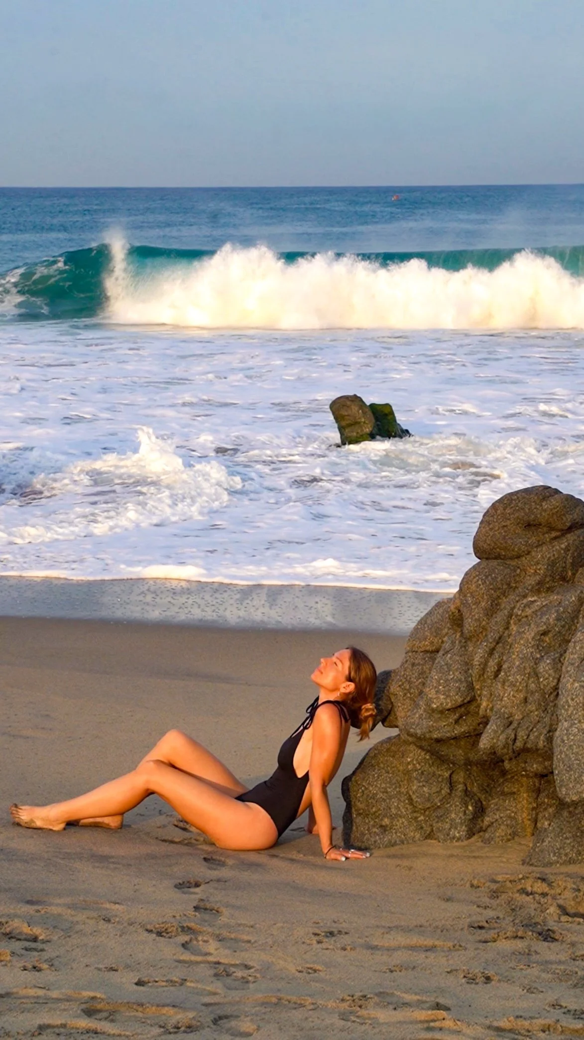 Woman in a black swimsuit sitting on the sandy beach, leaning against rocks, with waves crashing in the background.
