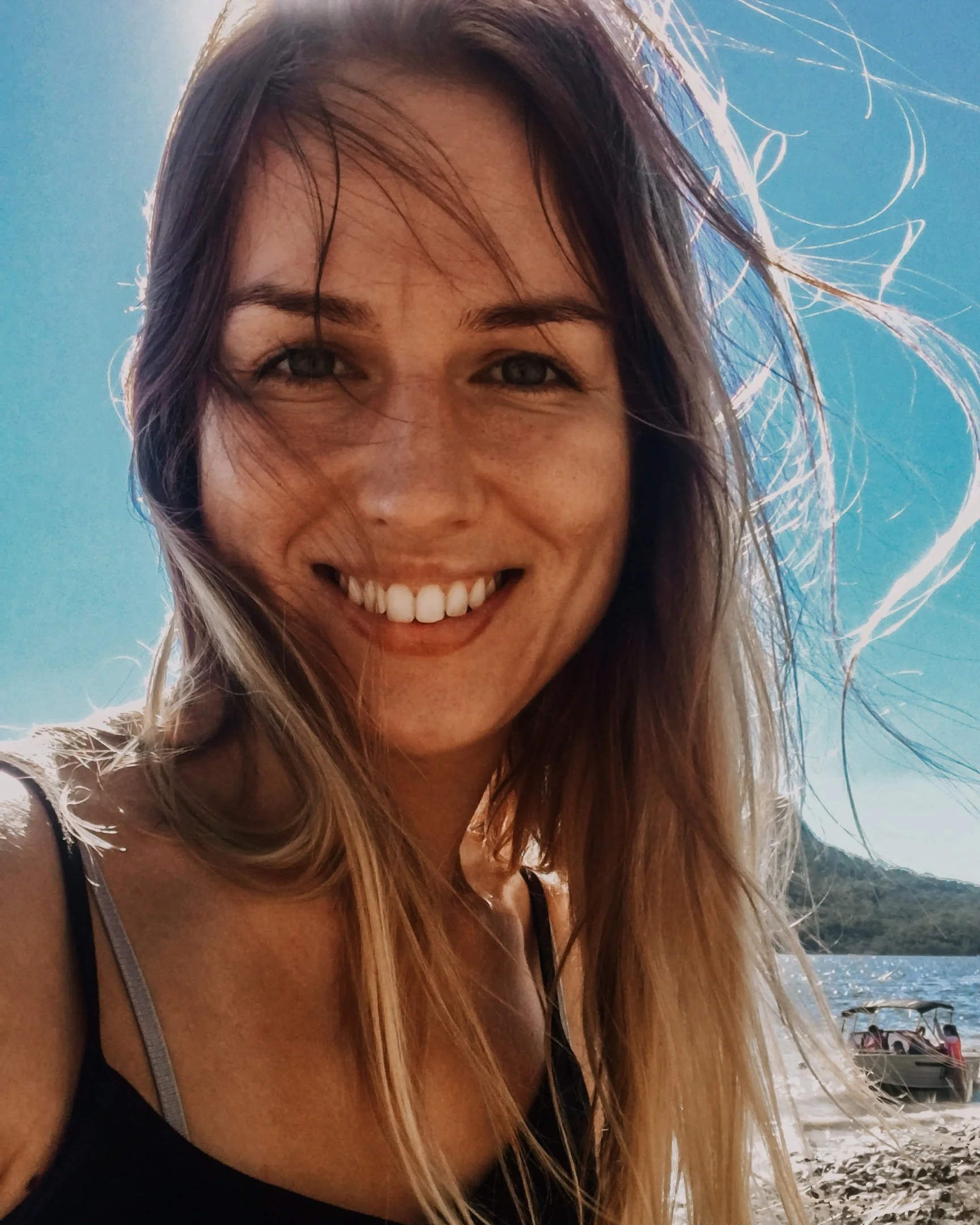 A smiling woman with long, windblown hair takes a selfie near a body of water and a boat, with a bright blue sky in the background.