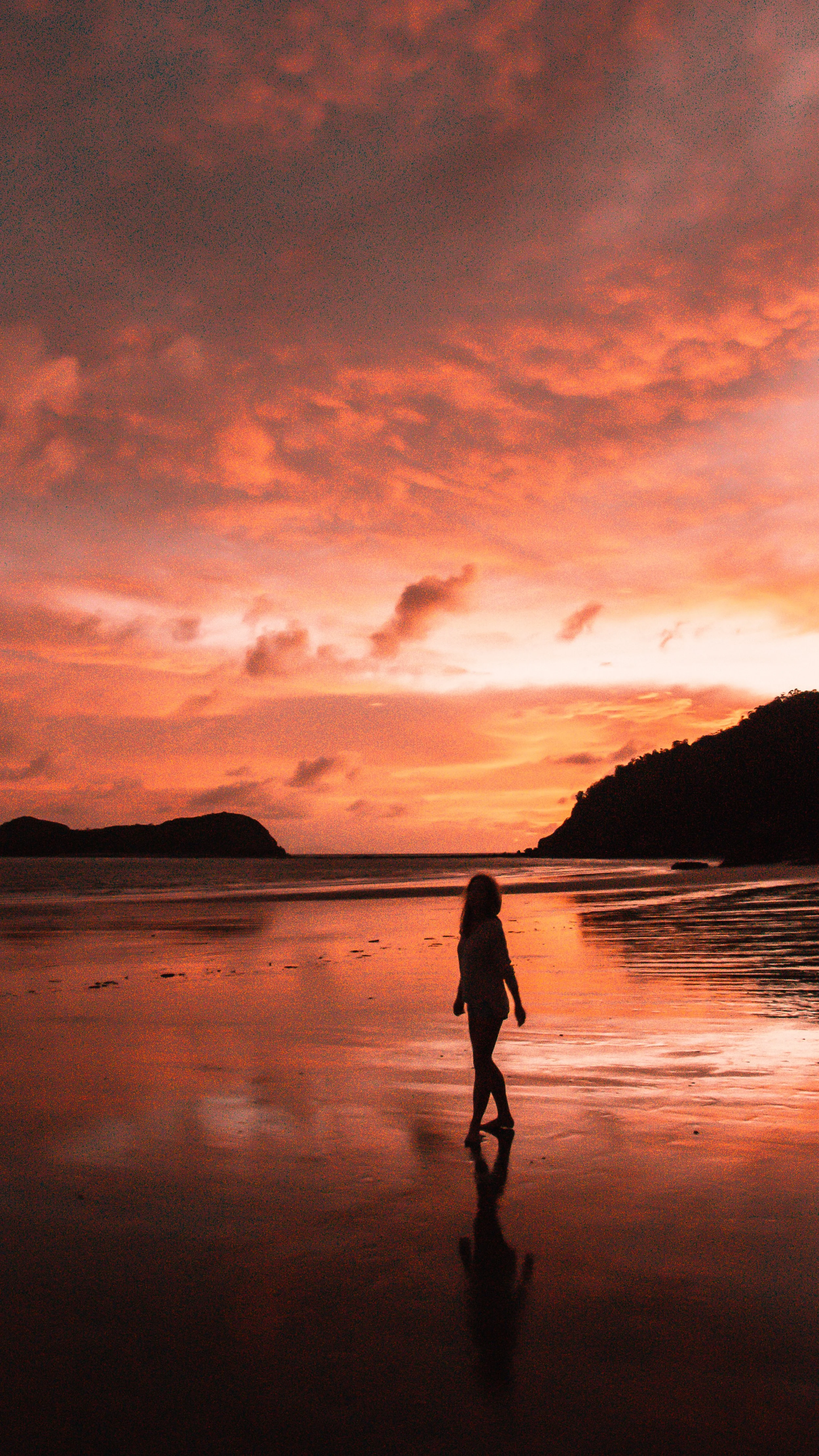 A person walking along a beach at sunset, with orange and pink clouds in the sky and the ocean in the background.