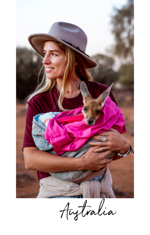A woman wearing a wide-brimmed gray hat and maroon shirt holding a kangaroo wrapped in a pink blanket outdoors.