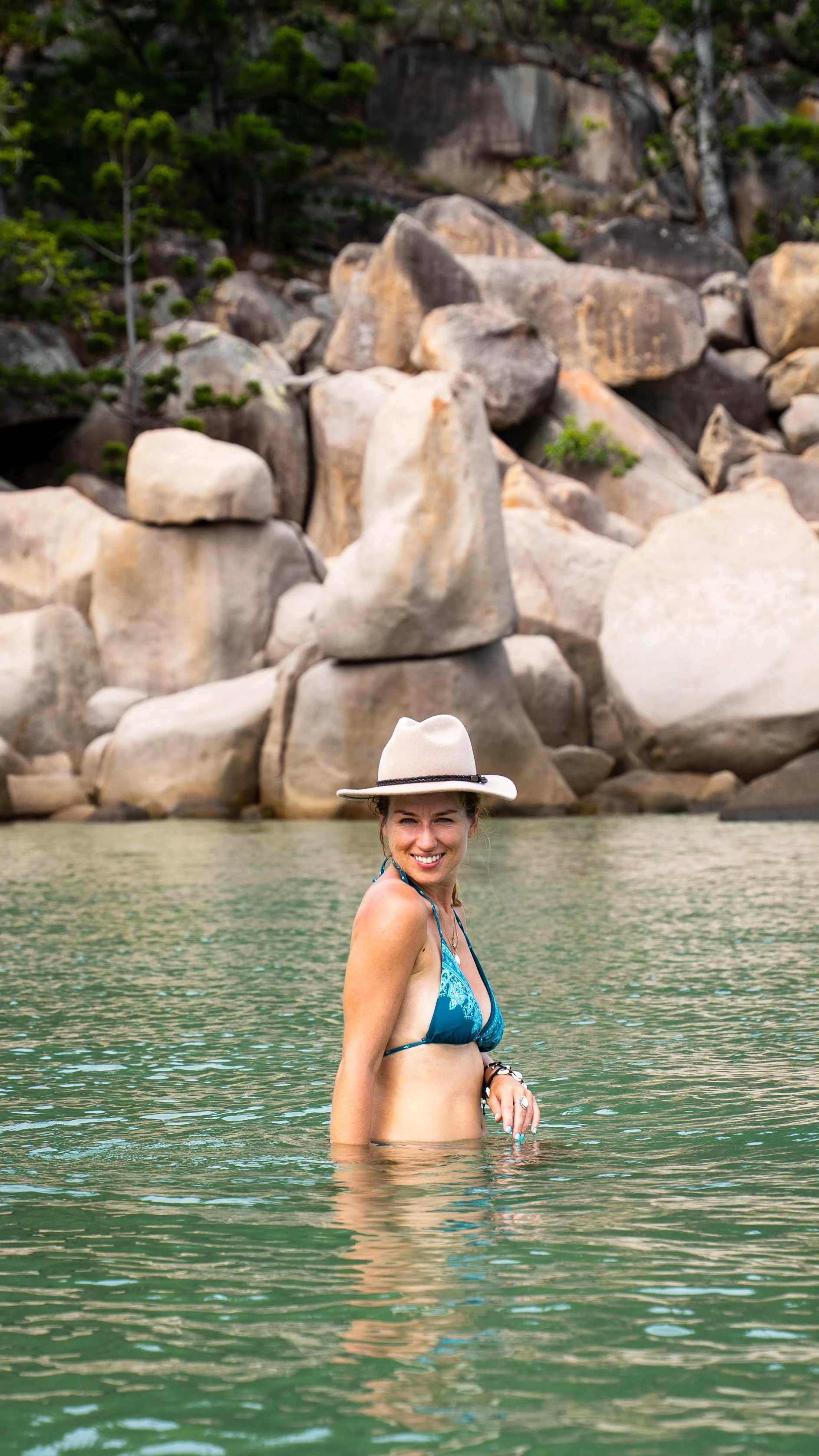 A woman smiling, wearing a blue bikini and a wide-brimmed hat, standing in a body of water with large rocks and green trees in the background.