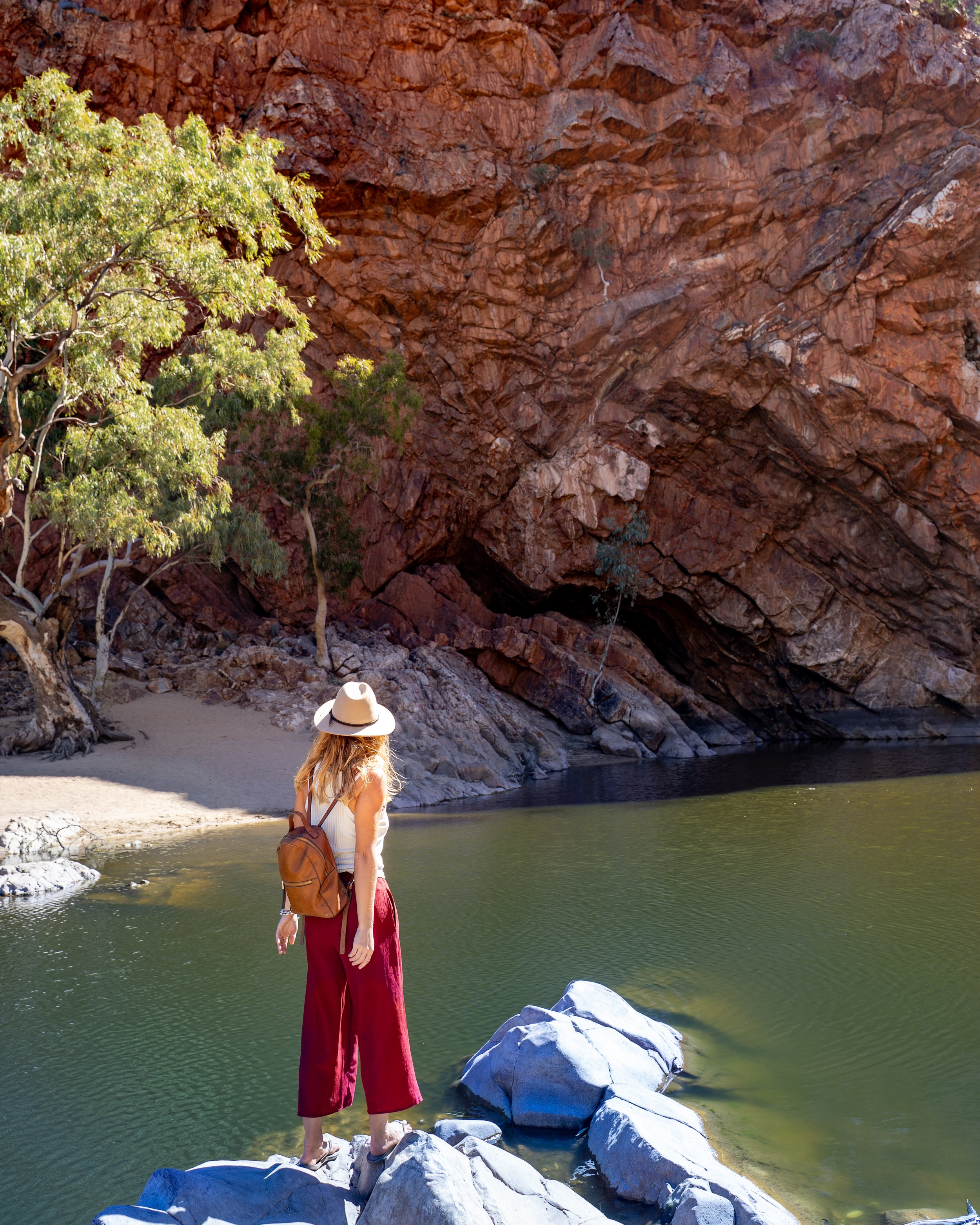 A woman with a hat and backpack standing on rocks by a river, with a large red rock formation and trees in the background.