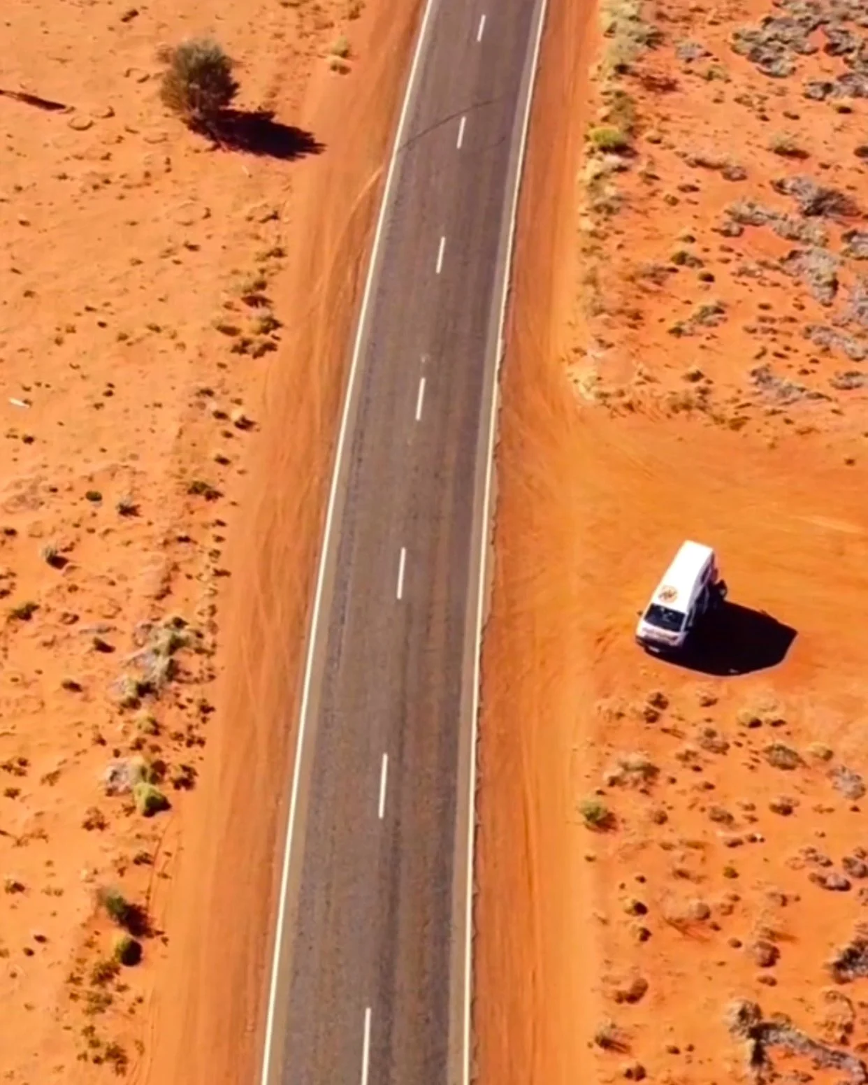 An aerial view of a deserted road with a small bus on the right side, surrounded by red desert terrain.