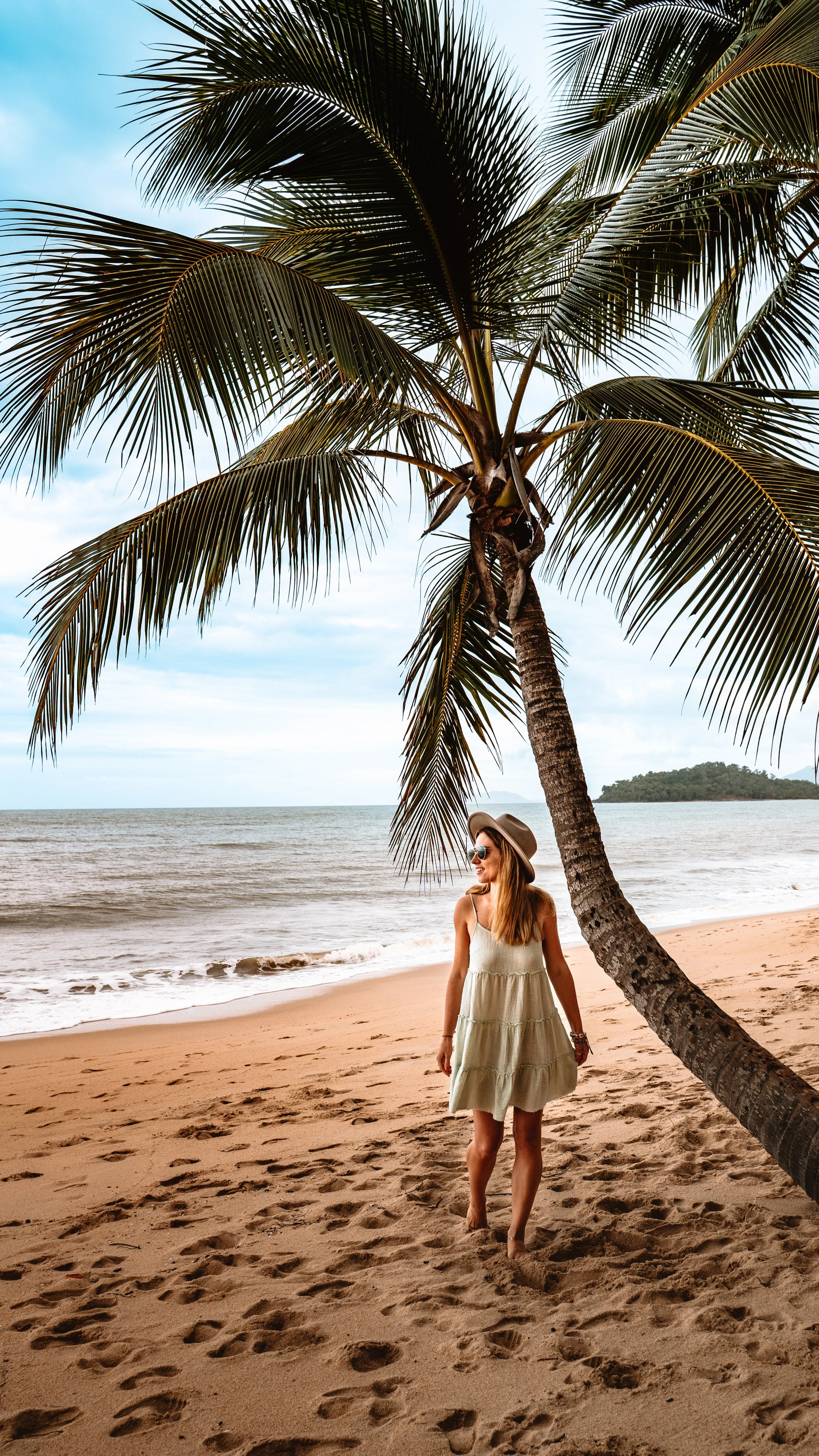 Woman walking on sandy beach under a tall palm tree, wearing a light-colored dress, wide-brimmed hat, and sunglasses, with ocean and distant land in the background.