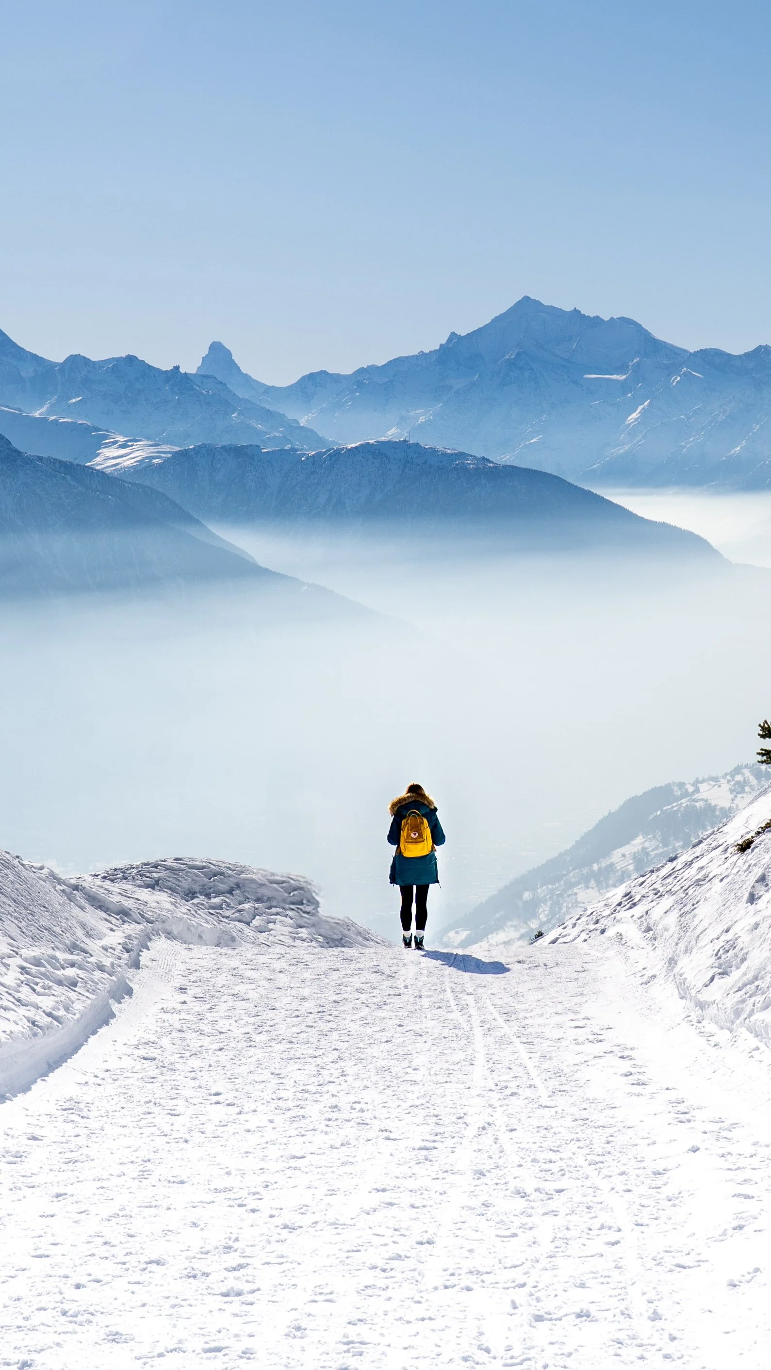A person with a yellow backpack standing on a snowy mountain trail, overlooking a valley and distant snow-capped mountains under a clear blue sky.
