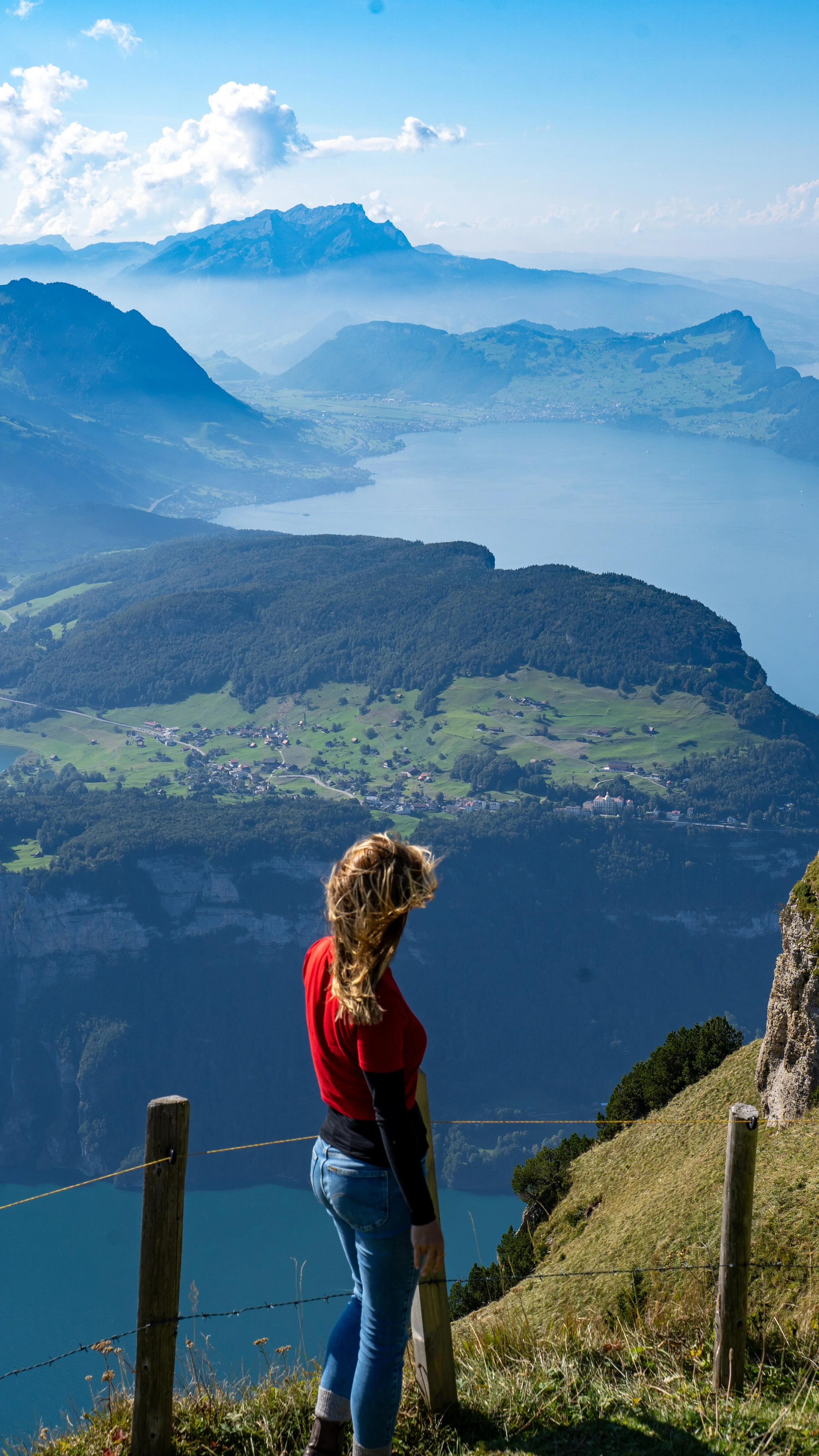 A woman standing on a hillside overlooking a large lake, mountains, and a picturesque landscape in the distance.