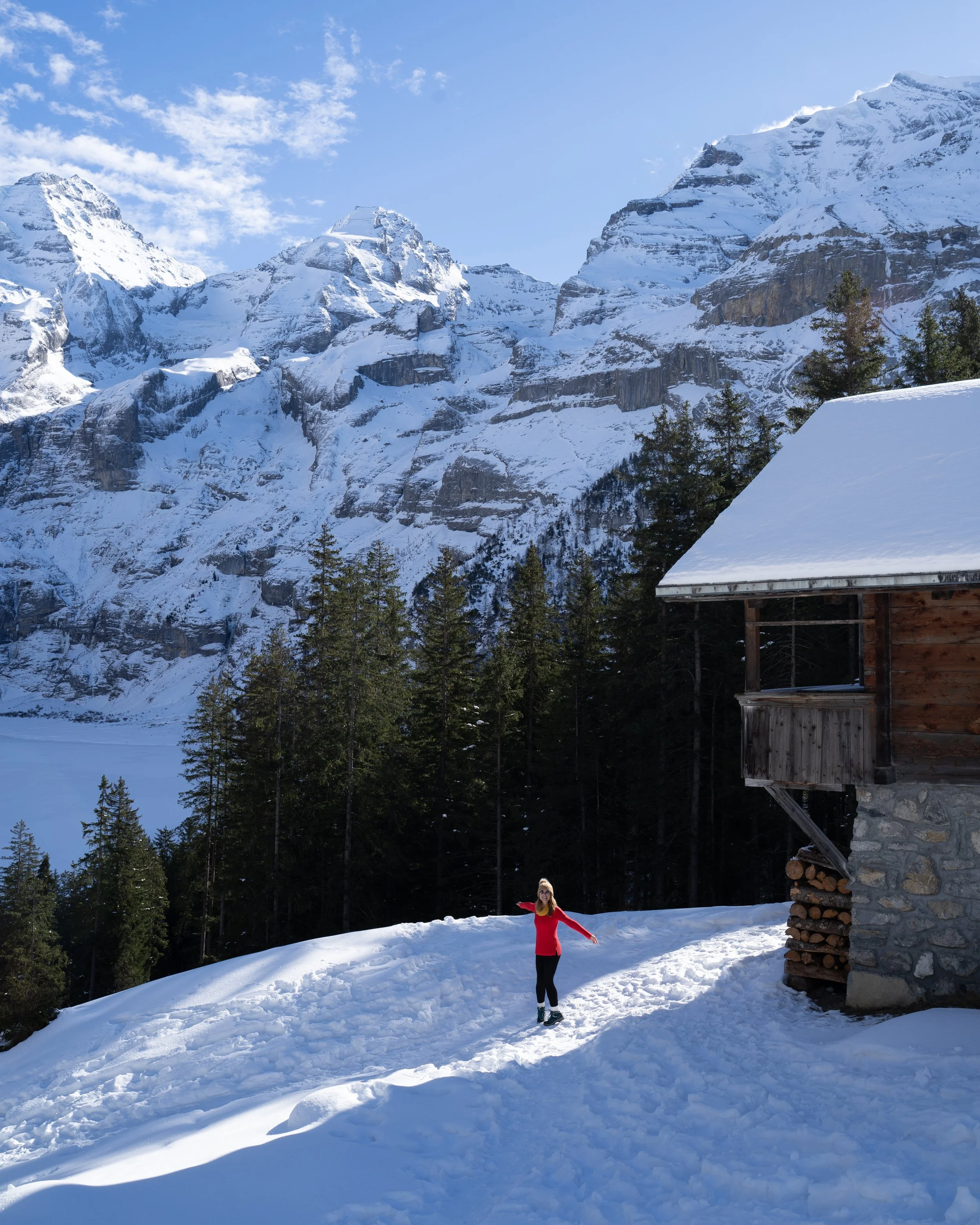A woman in a red jacket standing on snow-covered ground near a wooden cabin, with snow-capped mountains and a forest in the background.