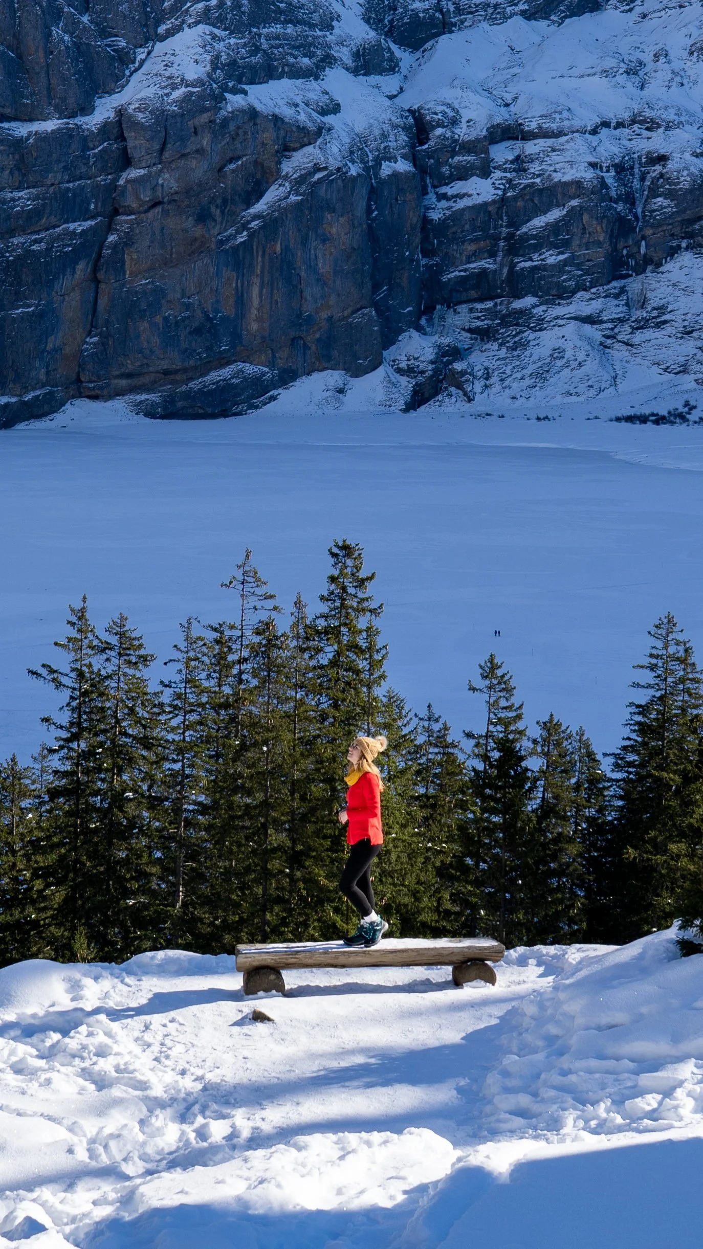 A woman in a red jacket and black pants walking on a wooden log bench in a snowy landscape with evergreen trees, a frozen lake, and tall rocky cliffs in the background.