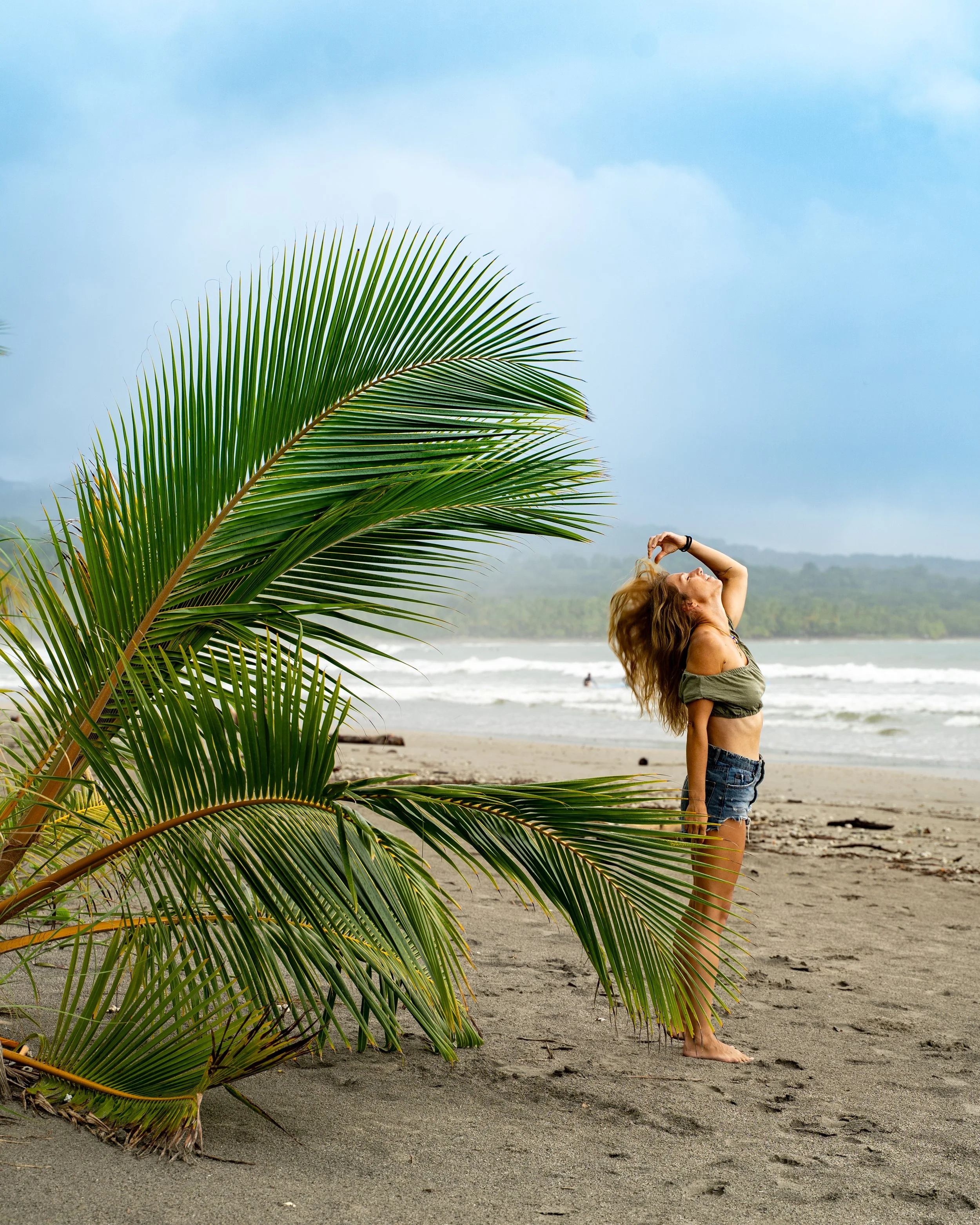 A woman with long, wavy hair stands on a beach, stretching with her hand on her forehead, near large green palm leaves, with the ocean and cloudy sky in the background.