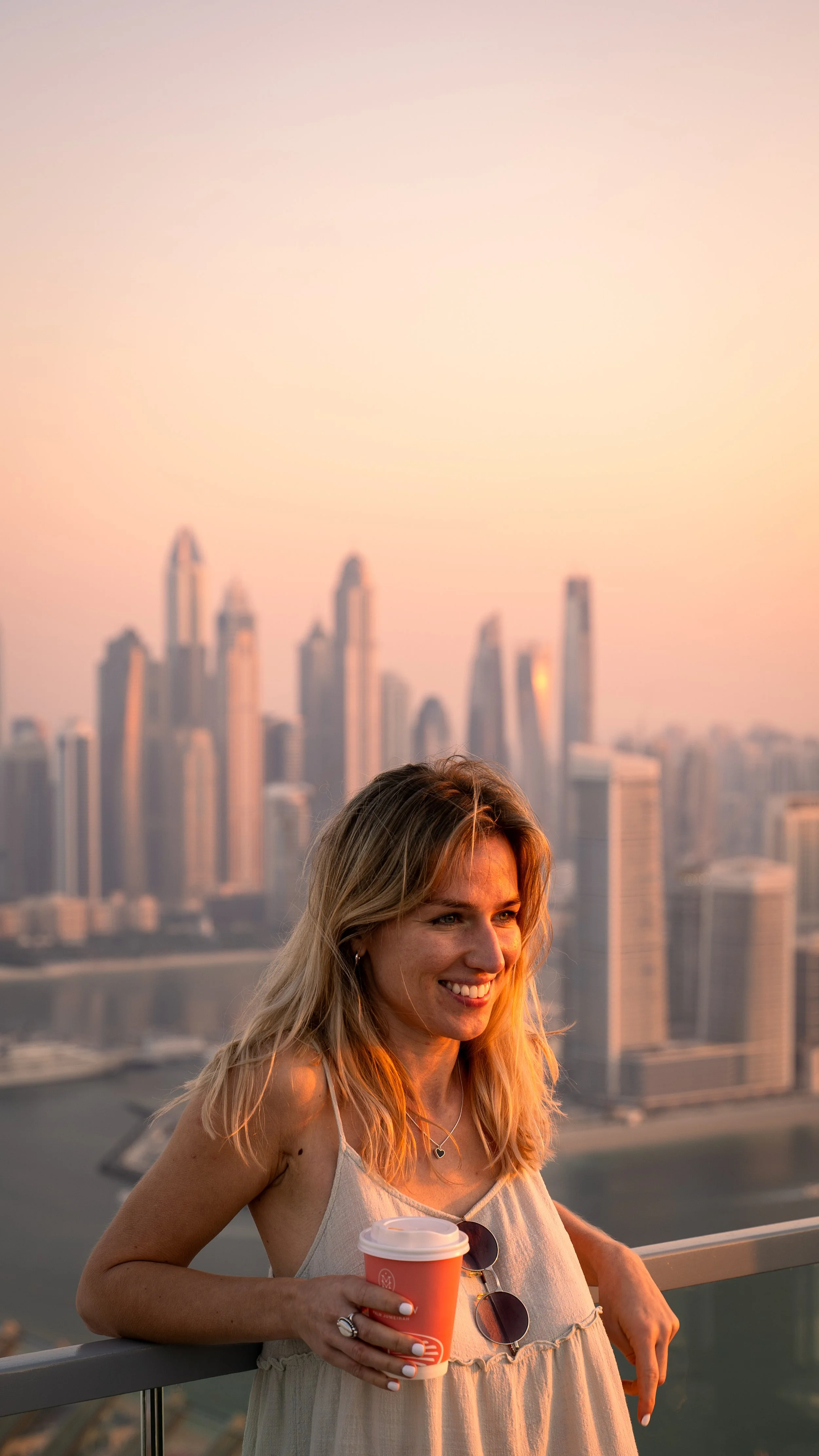 A woman smiling on a balcony holding a coffee cup, with a city skyline at sunset in the background.