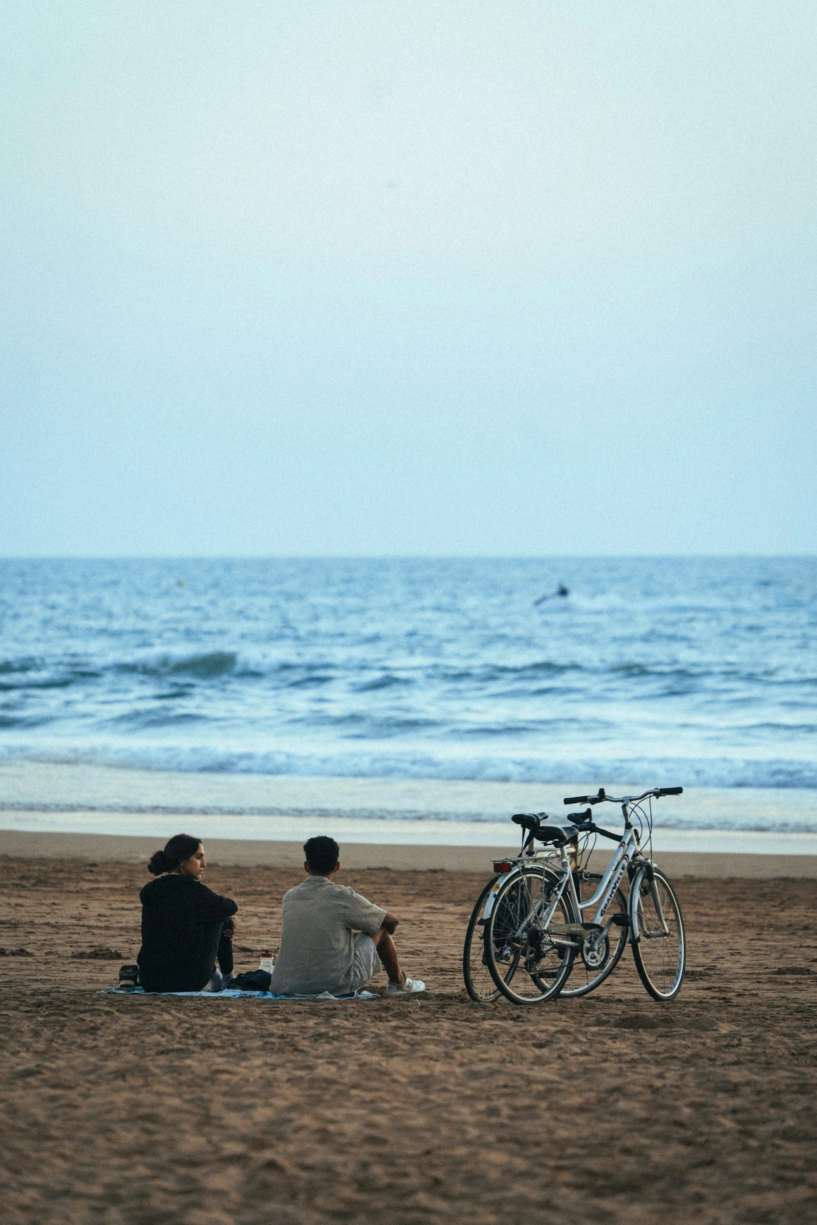 Two people peacefully sitting on a blanket on a beach near the ocean, with two bicycles parked next to them, and a boat visible in the distance on the water.