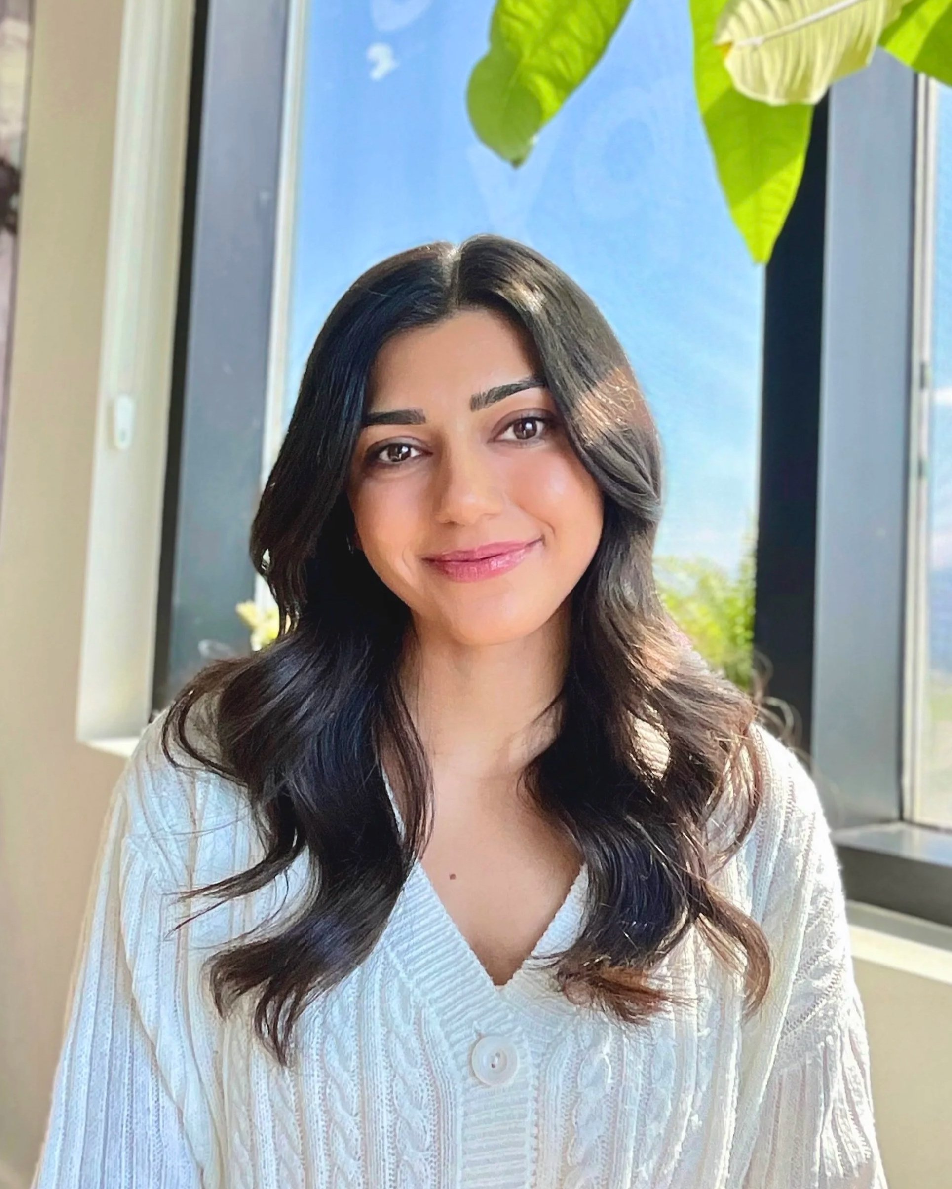 A woman with long, dark wavy hair smiling warmly and empathetically while seated indoors near a window with a blue sky and green leaves outside.