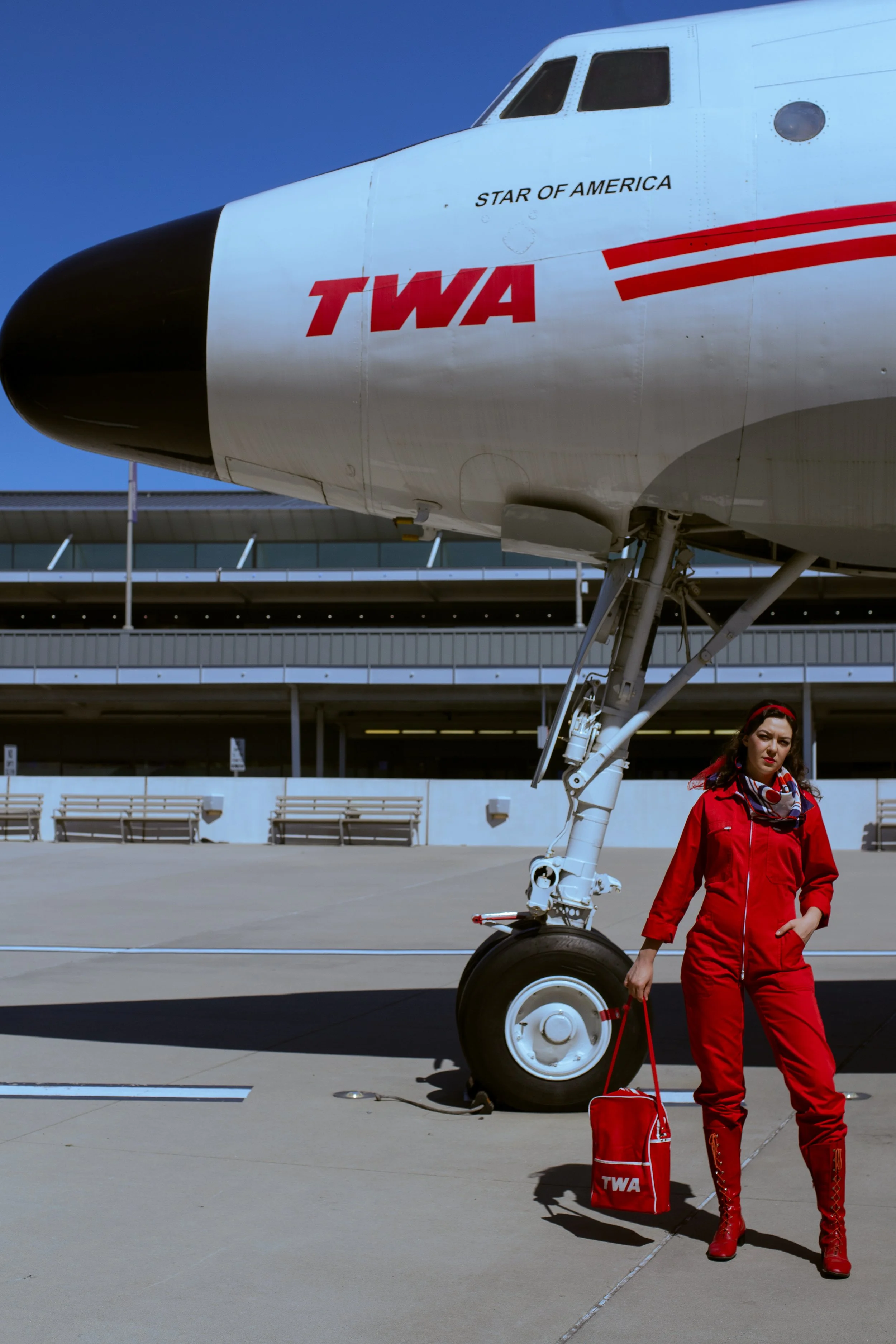 A woman dressed in a red jumpsuit, red boots, and a red cap, standing next to an airplane's nose with the text 'TWA' and 'Star of America' on it, at an airport.