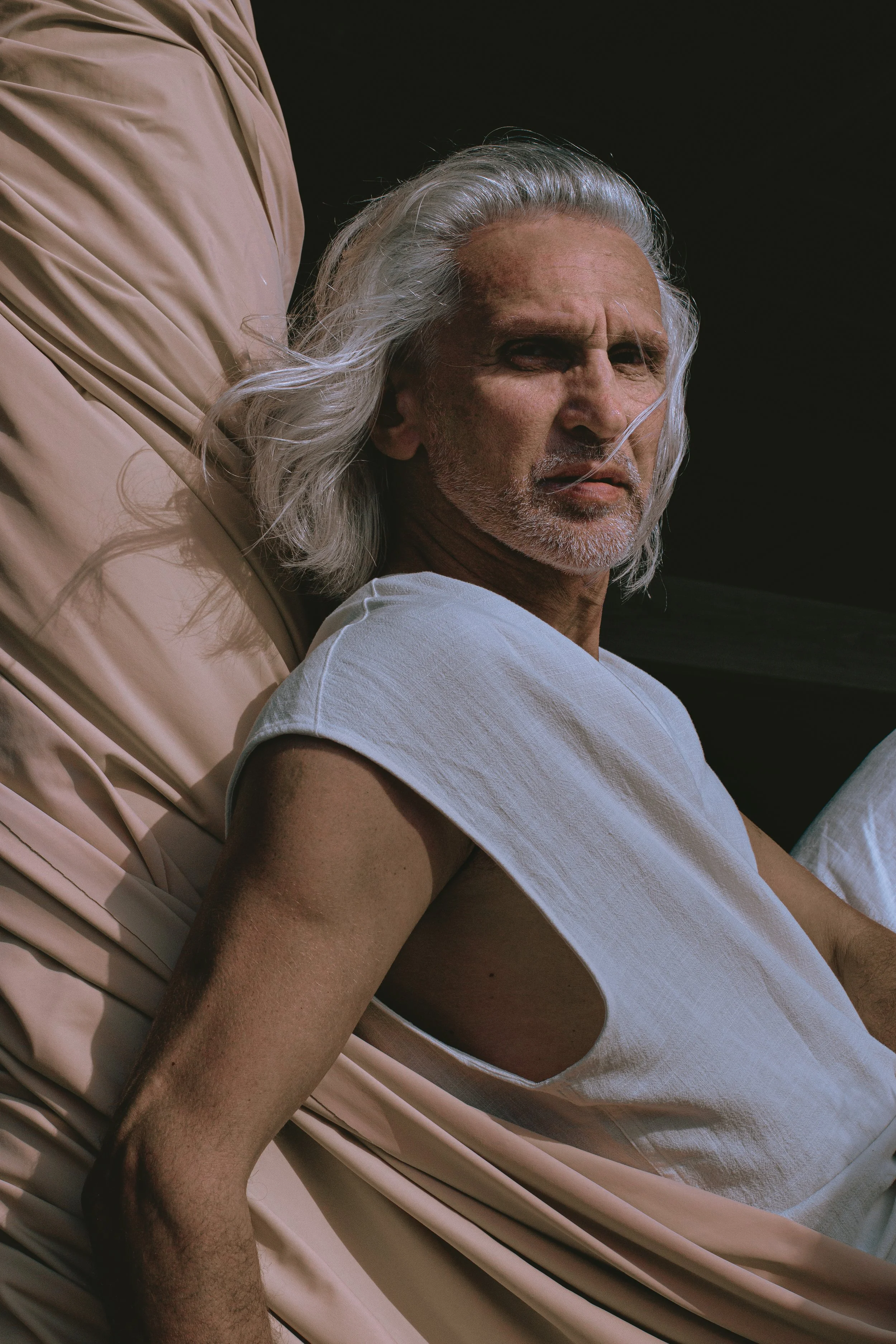 A man with long gray hair and a beard lying on a hospital bed with a beige sheet, wearing a white sleeveless shirt.