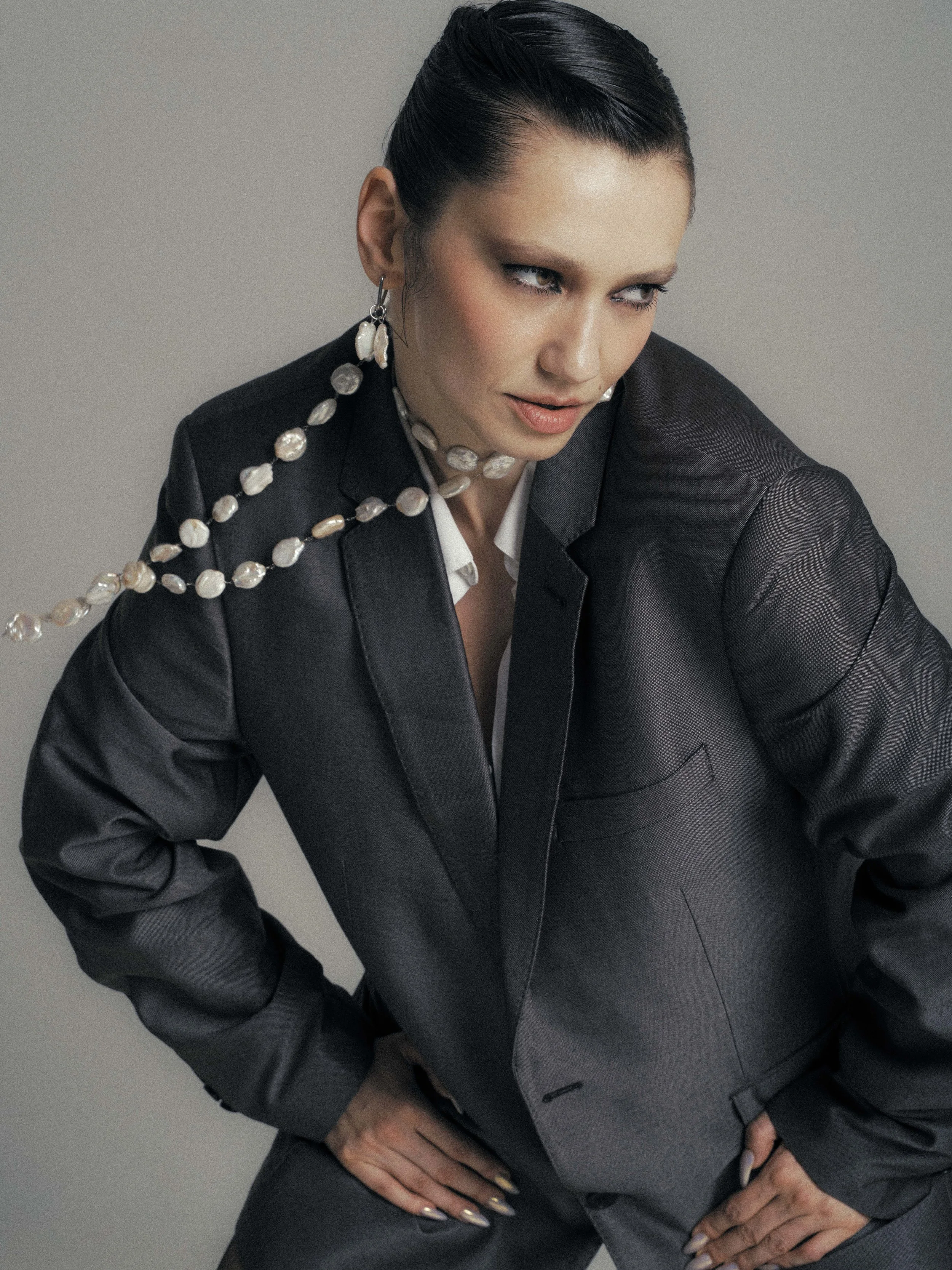 A woman with dark hair styled back, wearing a dark blazer over a light shirt, adorned with pearl jewelry, posing against a plain background.
