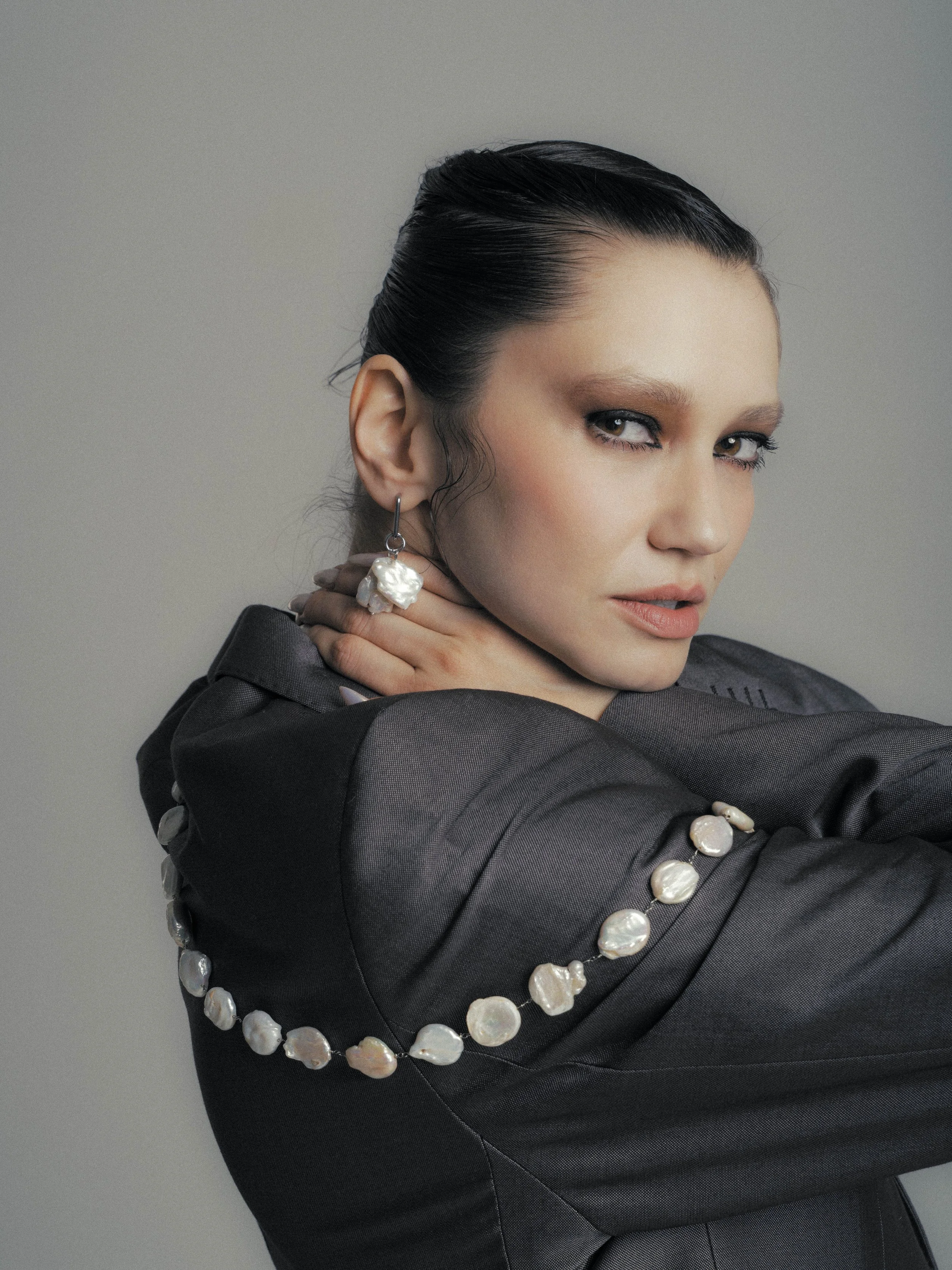 Close-up of a woman with dark hair in a slicked-back style, wearing a black blazer with pearl embellishments and large pearl earrings, posing against a plain gray background.