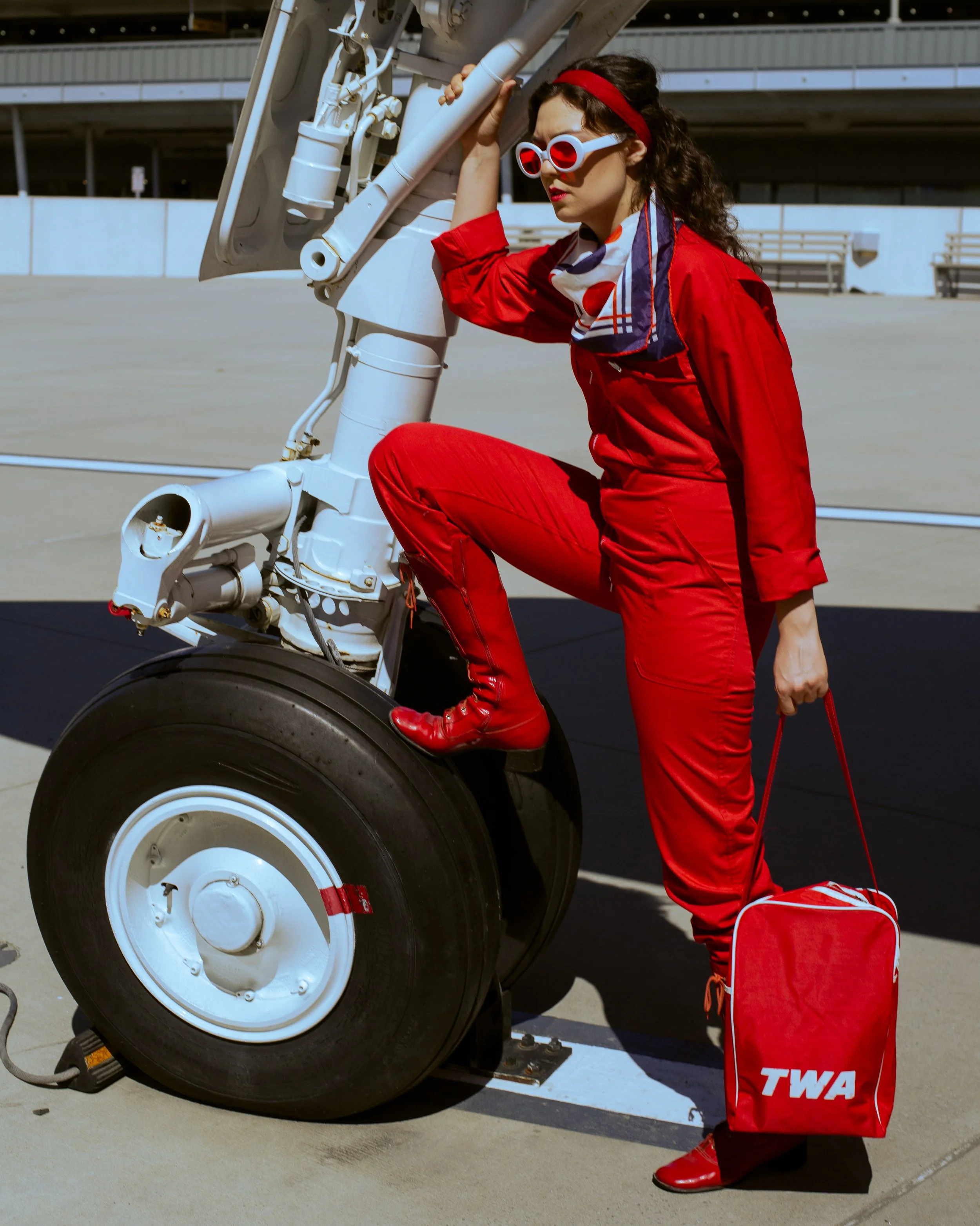 Woman in red jumpsuit, red boots, and red headband leaning on airplane landing gear at an airport, holding a red TWA trolley bag.