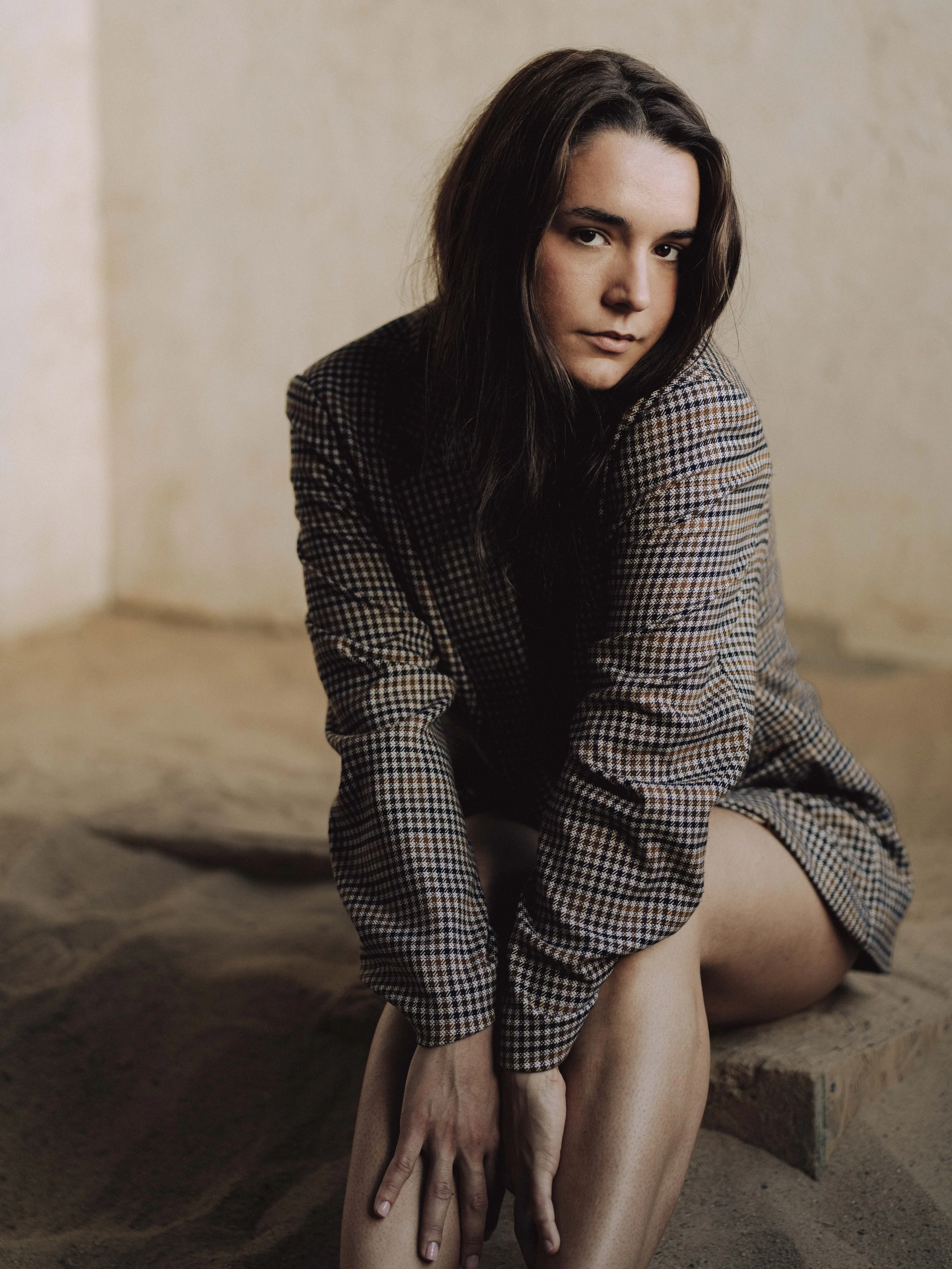 A woman with long dark hair sits on a sandy surface, wearing a plaid blazer and a matching skirt, with a neutral expression, looking toward the camera. Her left hand is resting on her knee, and her right arm is slightly bent.