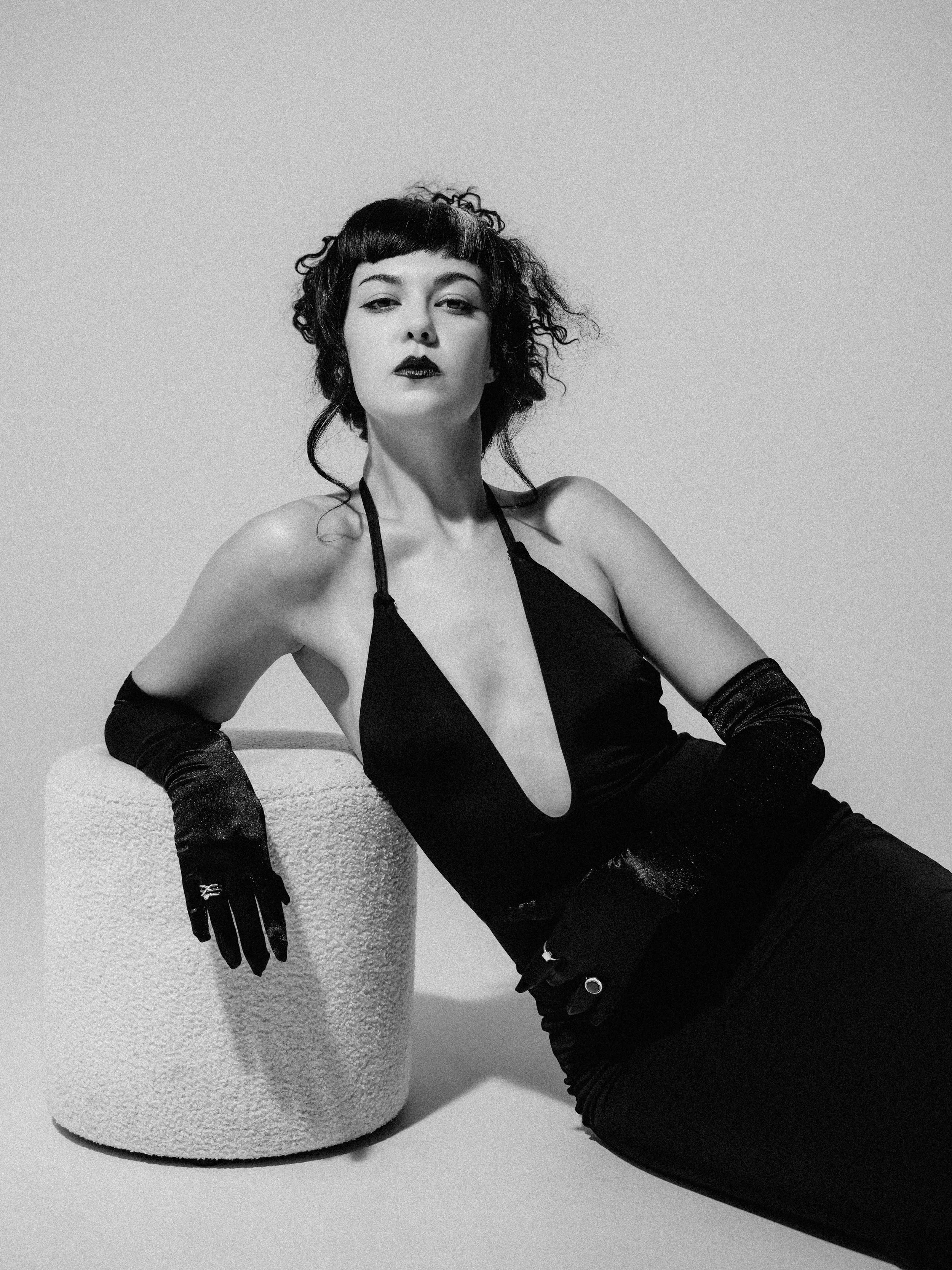 Black and white photo of a woman with short curly hair, wearing a spaghetti strap top and long gloves, sitting on the floor with a textured object behind her.