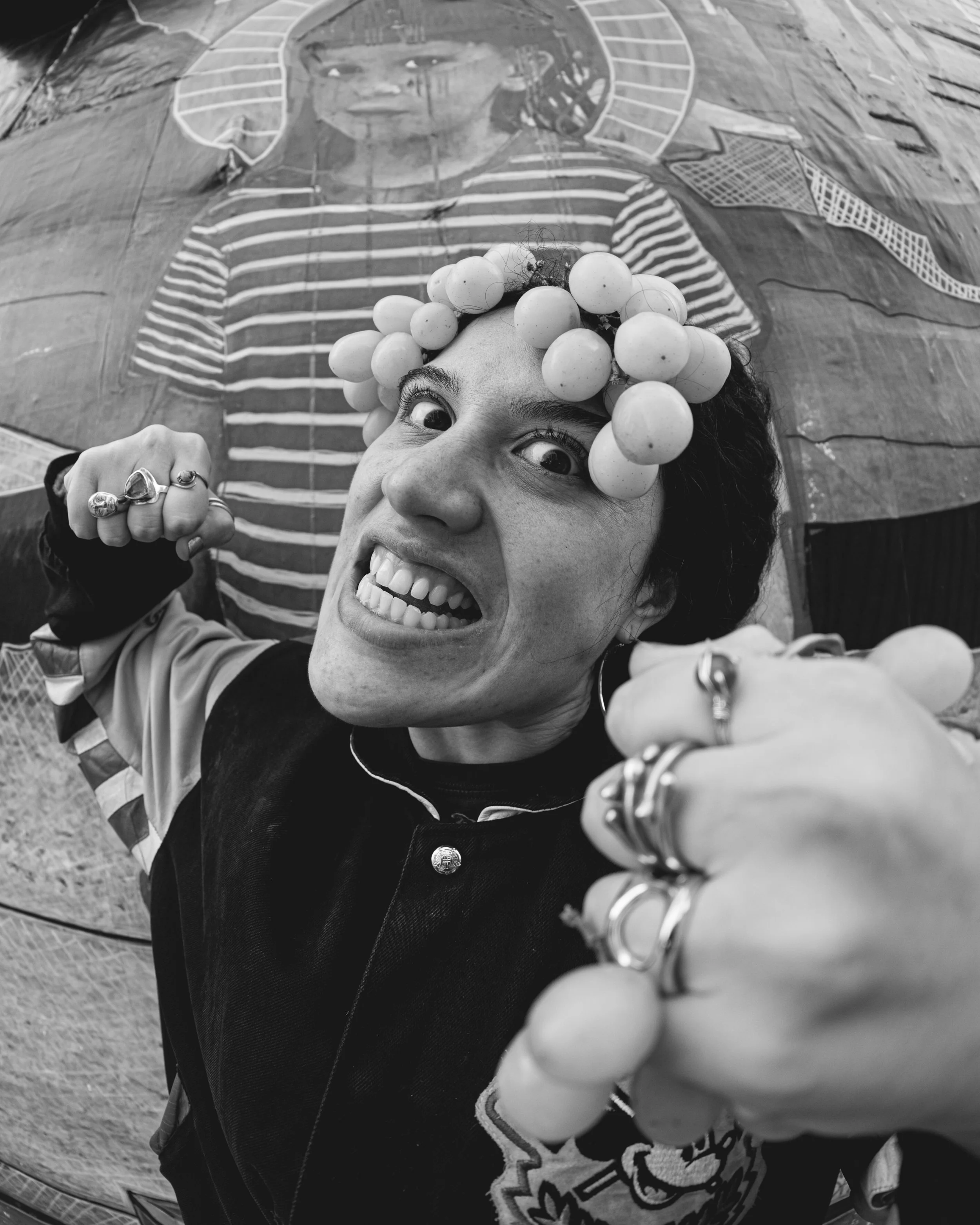 A woman with curly hair and multiple rings on her fingers is smiling at the camera, wearing a headband made of grapes, with a mural of a woman in striped shirt in the background.