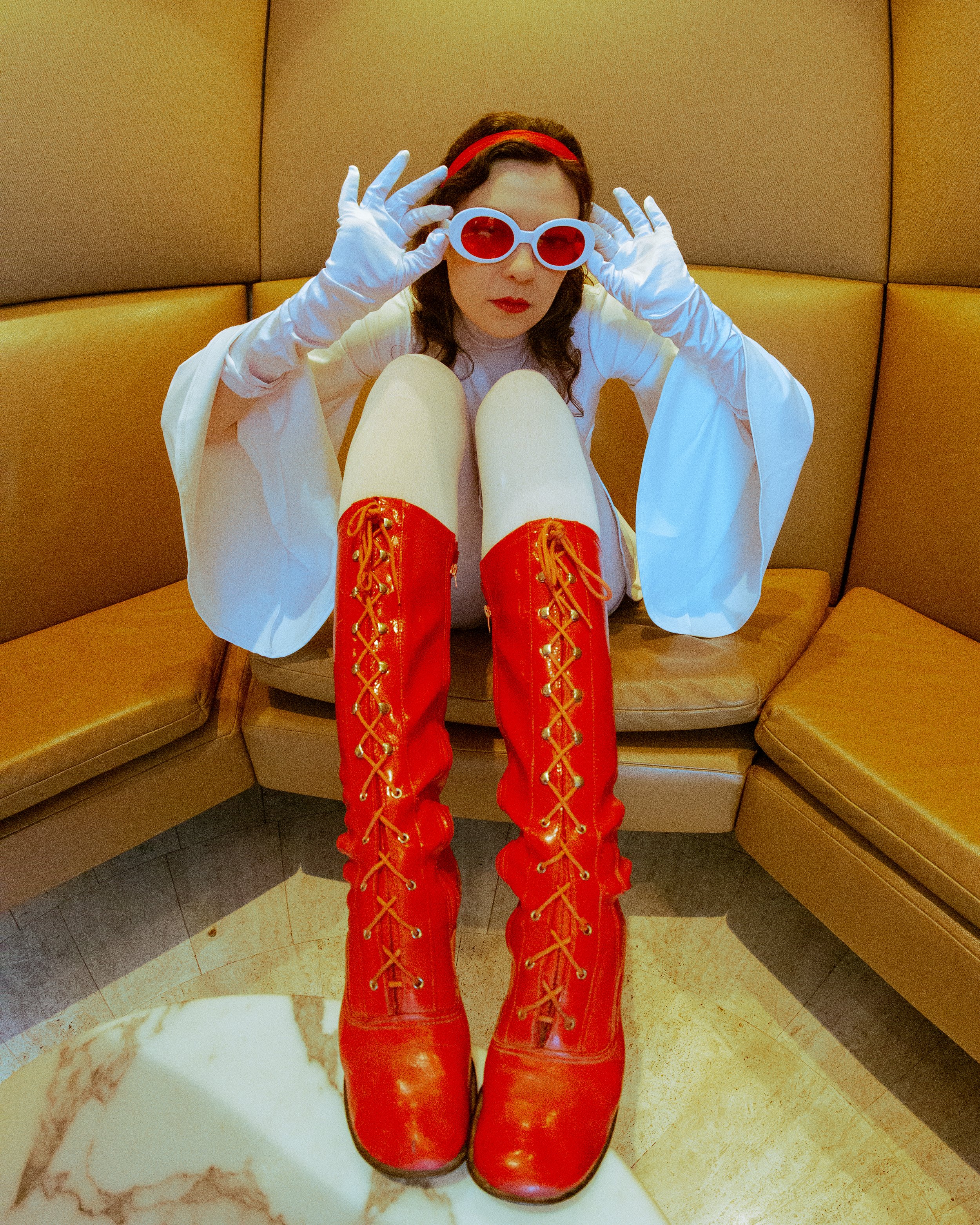 Person sitting on a yellow booth wearing red shiny lace-up boots, white gloves, white dress, red headband, and red sunglasses, with legs up and looking at the camera in a close-up shot.