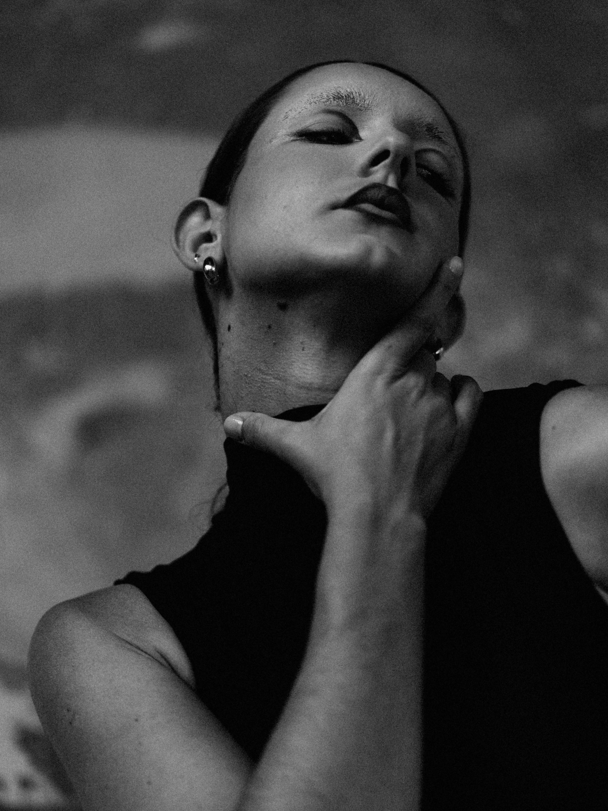 A black and white photo of a woman with dark lipstick and earrings, touching her neck with one hand, with her head tilted back slightly and a confident expression.
