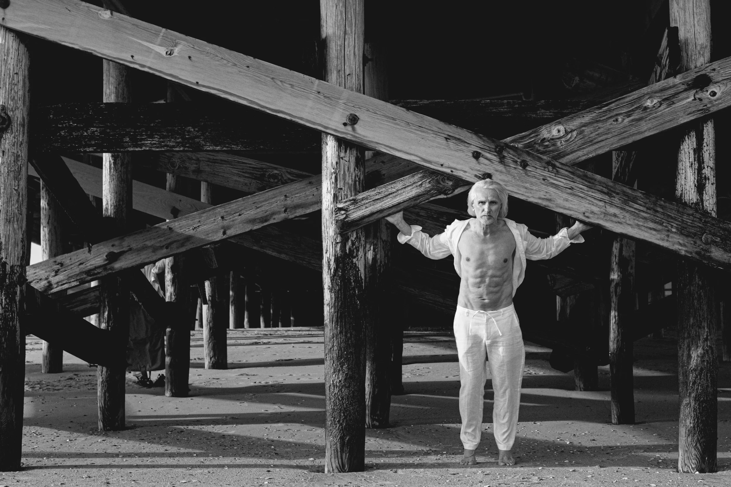 A shirtless elderly man with long white hair and a beard, standing under a wooden pier on the beach, holding onto the pier's beams, looking at the camera.