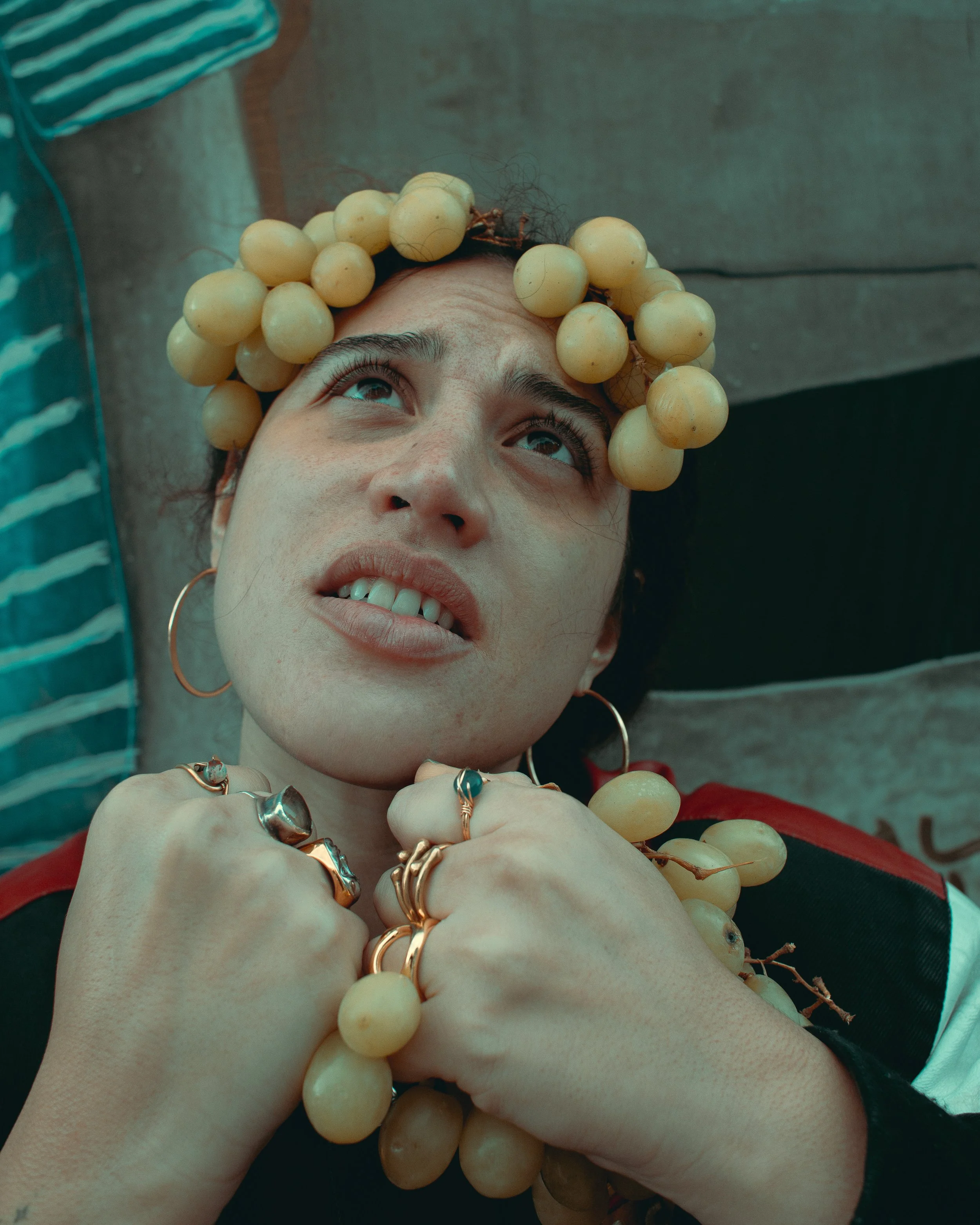 A person wearing a grape-shaped headpiece and jewelry, lying on a surface, with their head tilted back and looking upward.