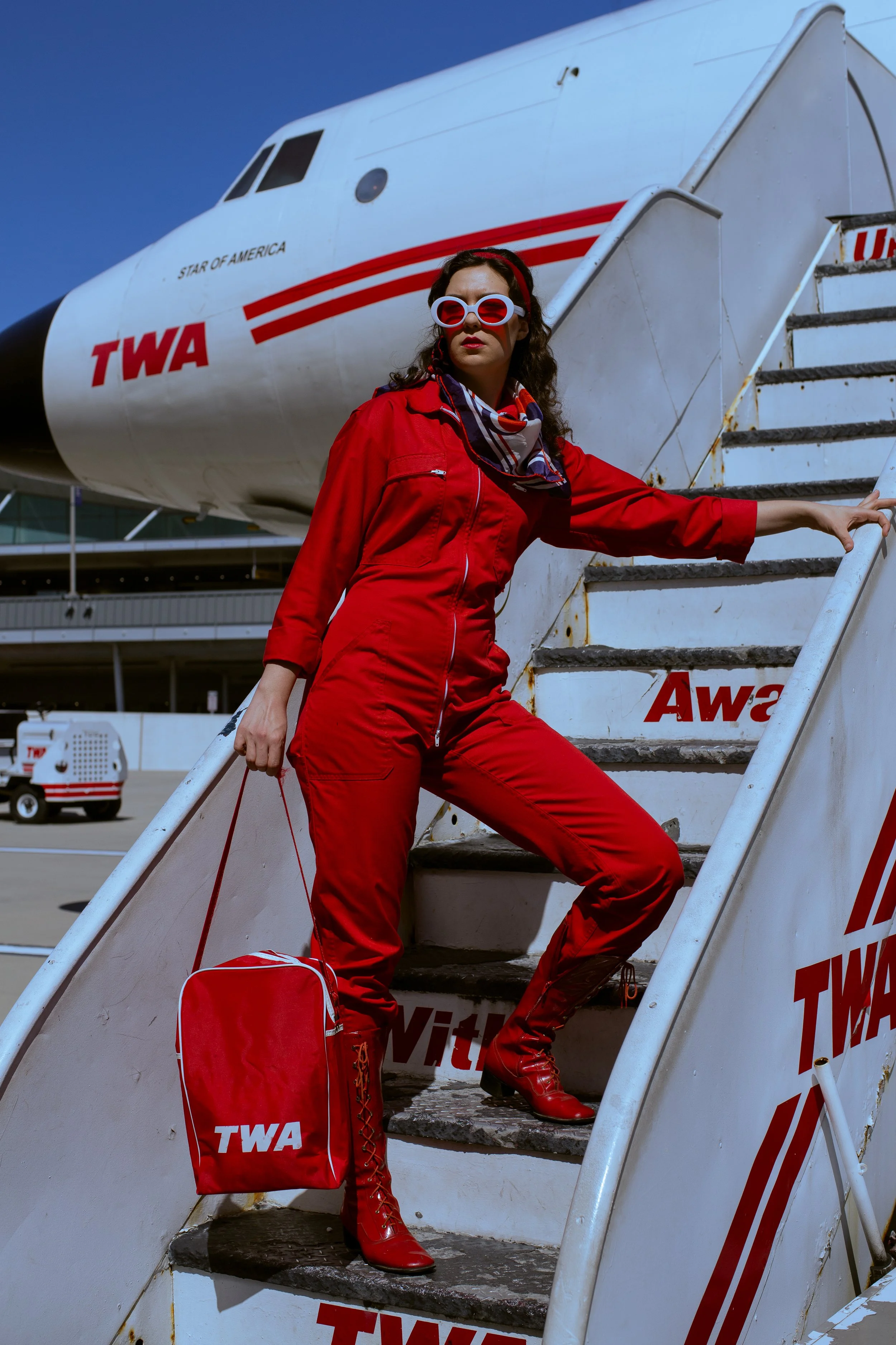 A woman dressed in a red jumpsuit and red boots, standing on the stairs of an airplane stairway with stairs and a luggage bag with TWA logo, at an airport.
