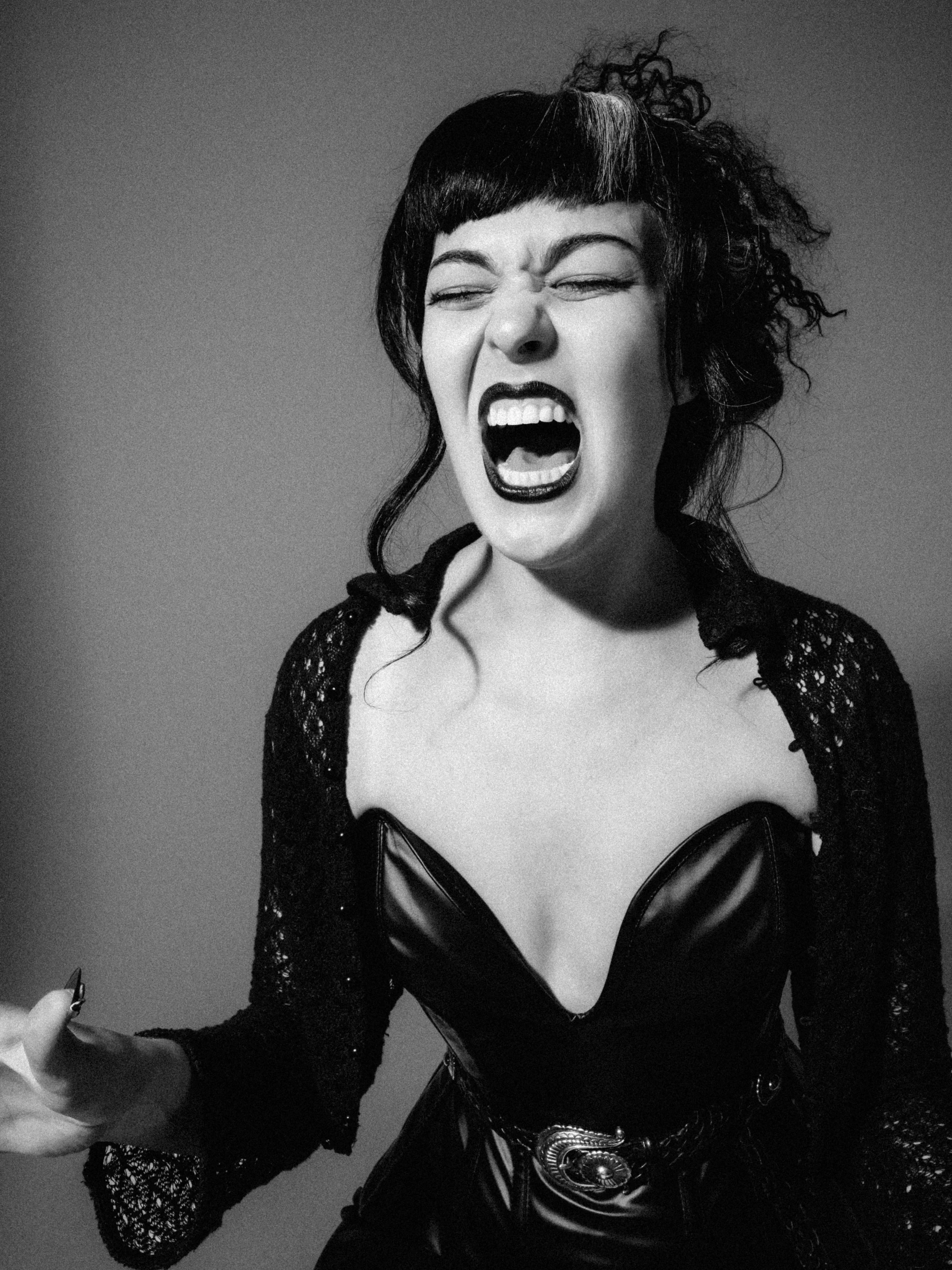 A woman with curly hair and bangs, wearing a black leather bustier and a lace cover-up, posing with her mouth open in a scream or yell.