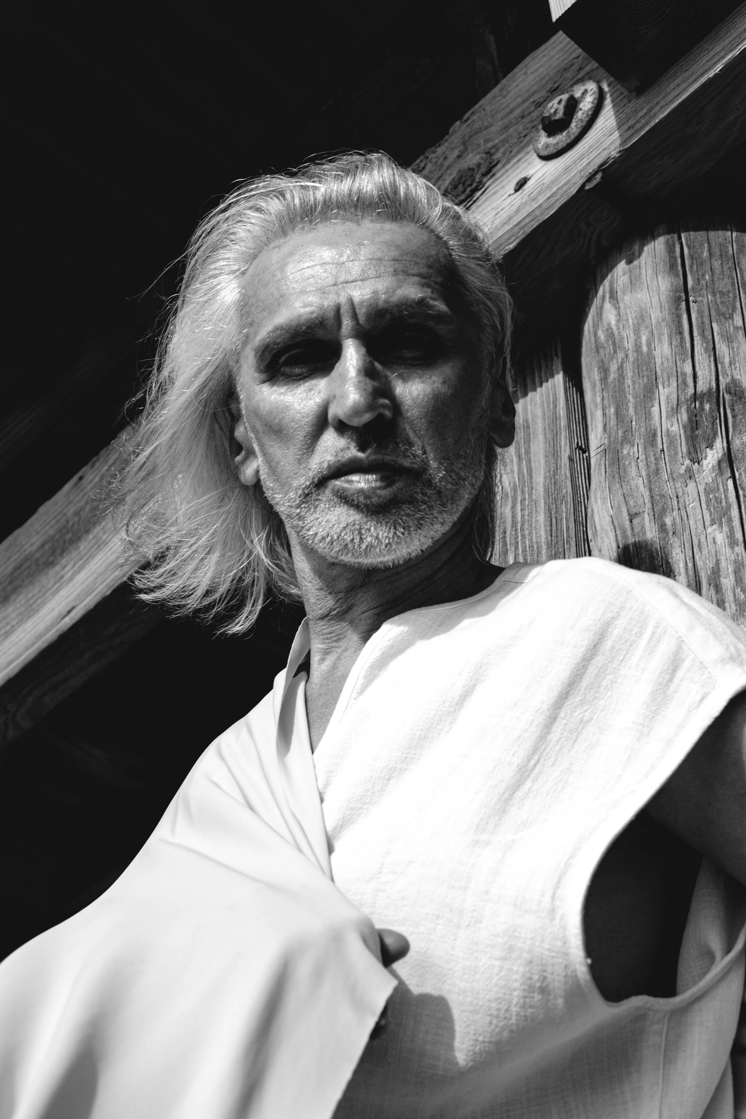 Close-up of a middle-aged man with long, wavy, gray hair, wearing a white shirt, standing outdoors against a wooden fence with a distinctive symbol or marking on a piece of wood, in black and white.