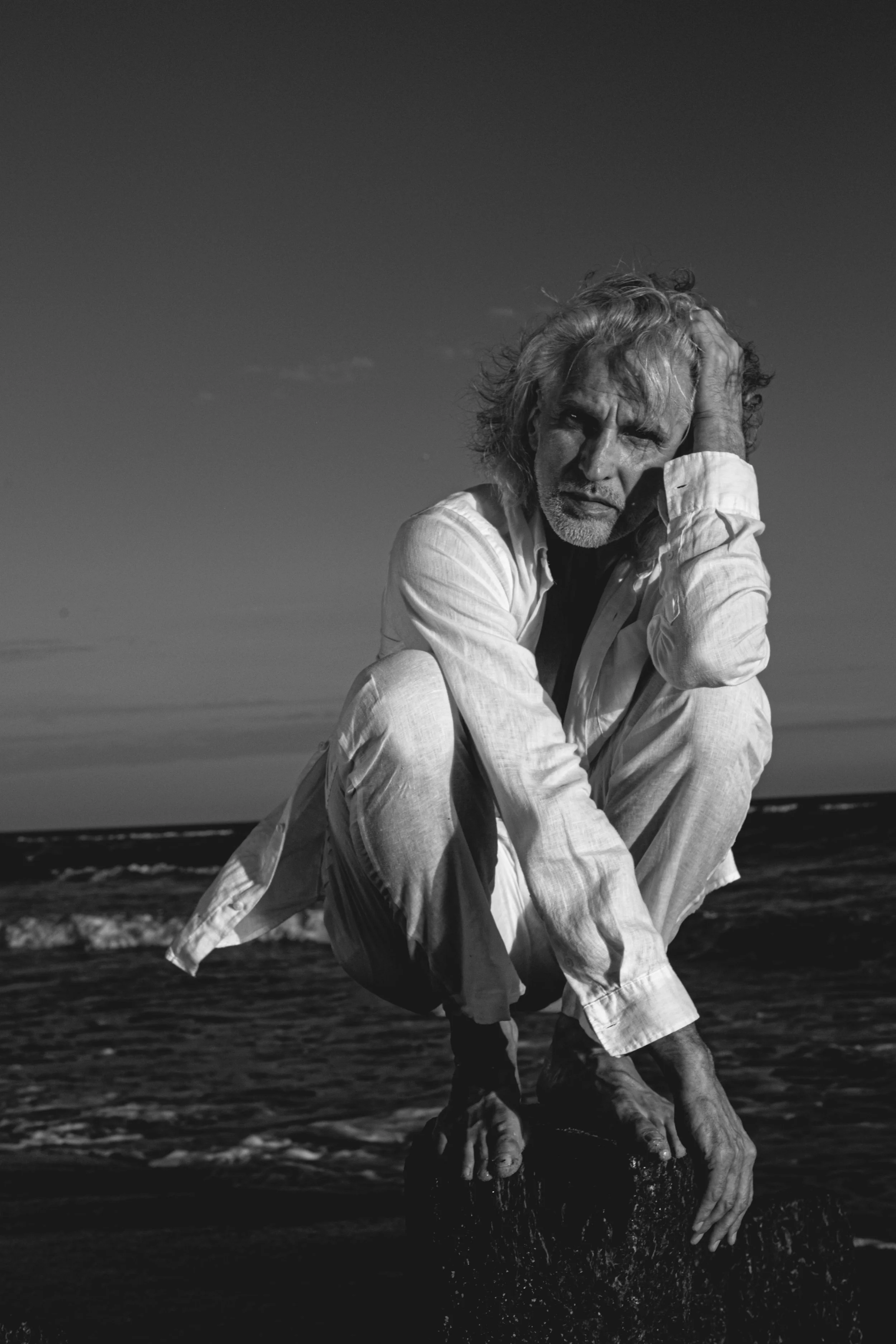 A black and white photo of a man with curly hair and a beard, sitting on a rock at the beach, with the ocean and sky in the background, holding his head with one hand, appearing contemplative.