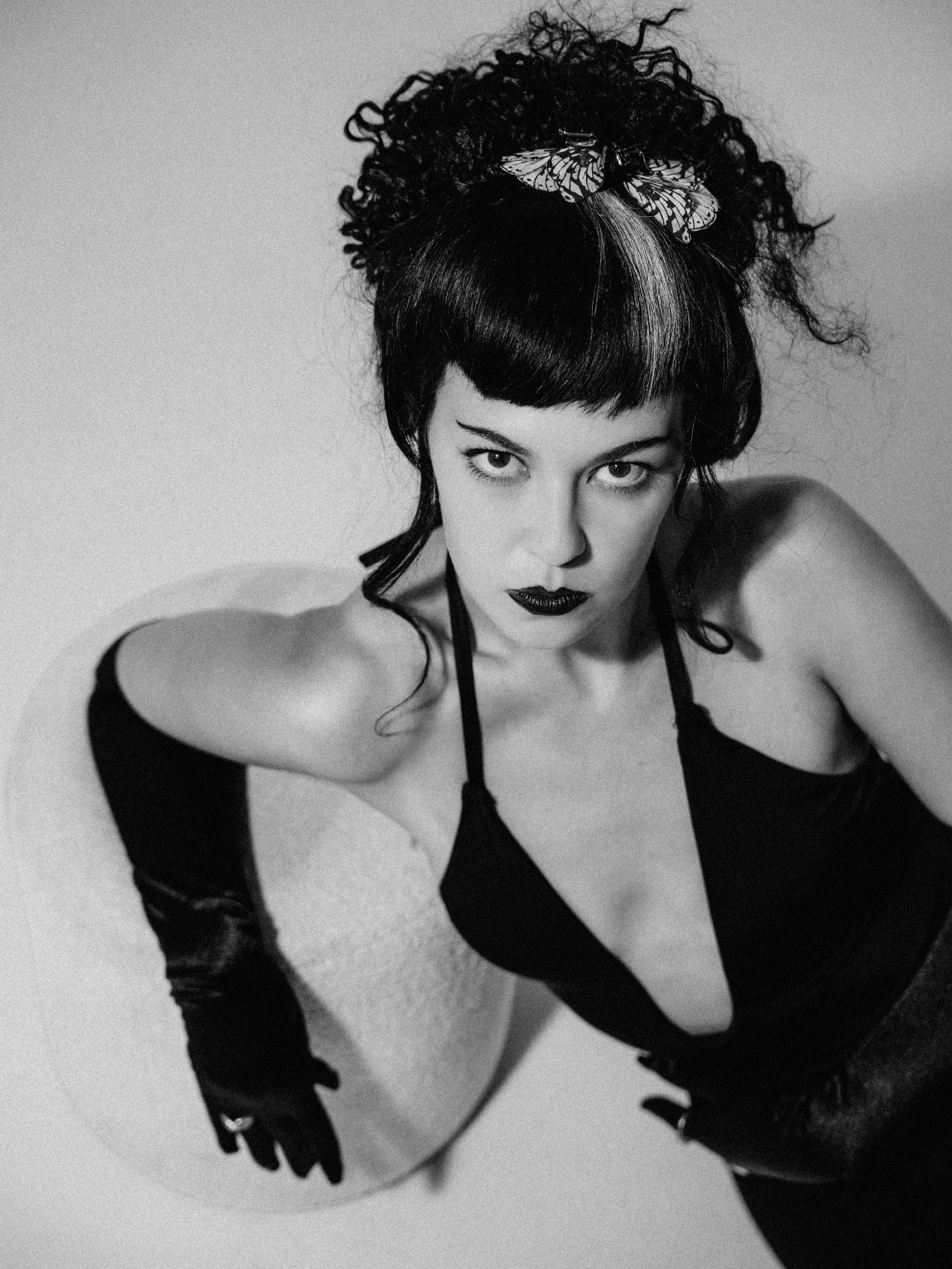 Black and white photograph of a woman with dark hair, styled with a butterfly hair clip, wearing dark lipstick, and a strappy black top, seated with her arms resting on her knees in front of a plain background.