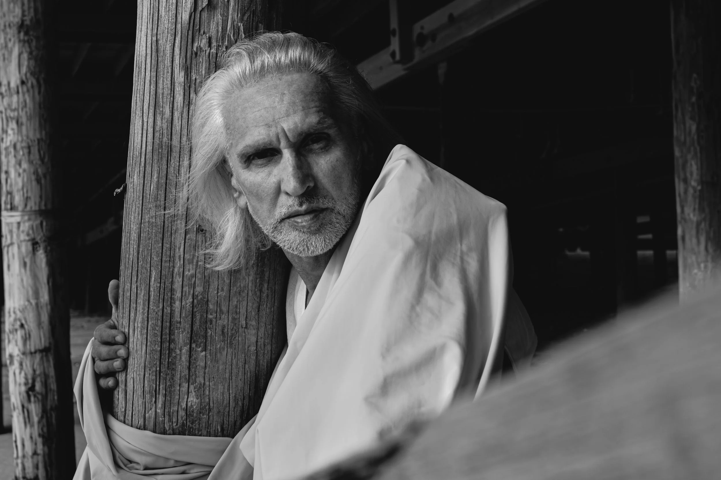 A black and white photo of an older man with long gray hair and a beard, leaning against a wooden post, wearing a light-colored shirt, outdoors, with a wooden structure in the background.