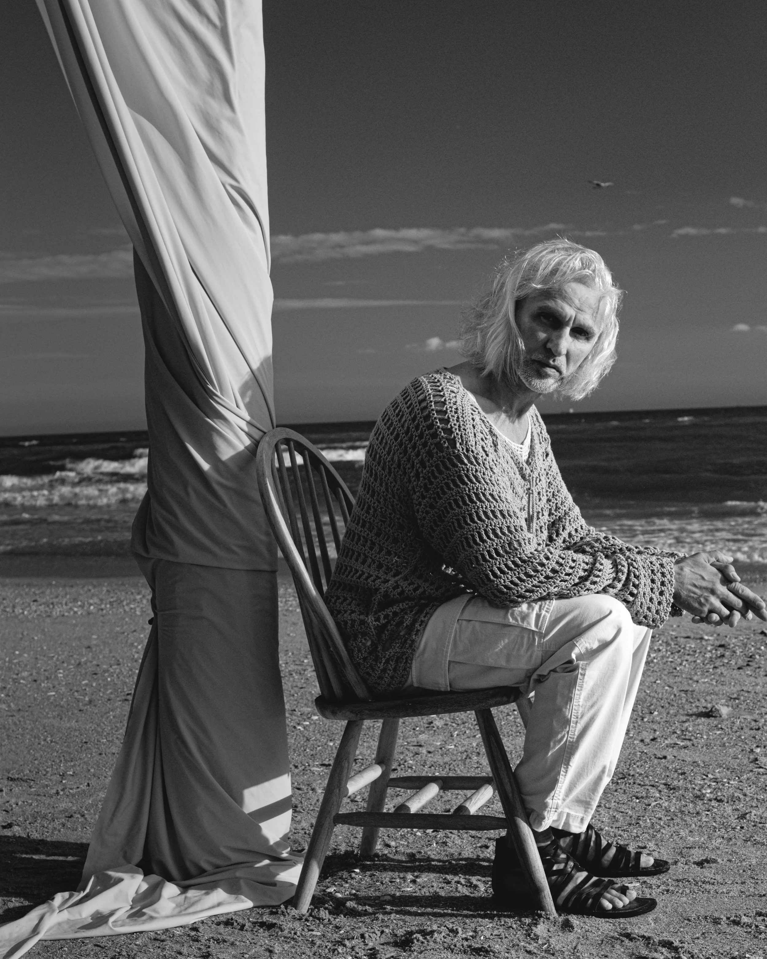 An elderly man with long gray hair and a beard sitting on a wooden chair on the beach, wearing a knitted sweater and light-colored pants, looking directly at the camera. Next to him, part of another person is visible, standing and partly covered by a