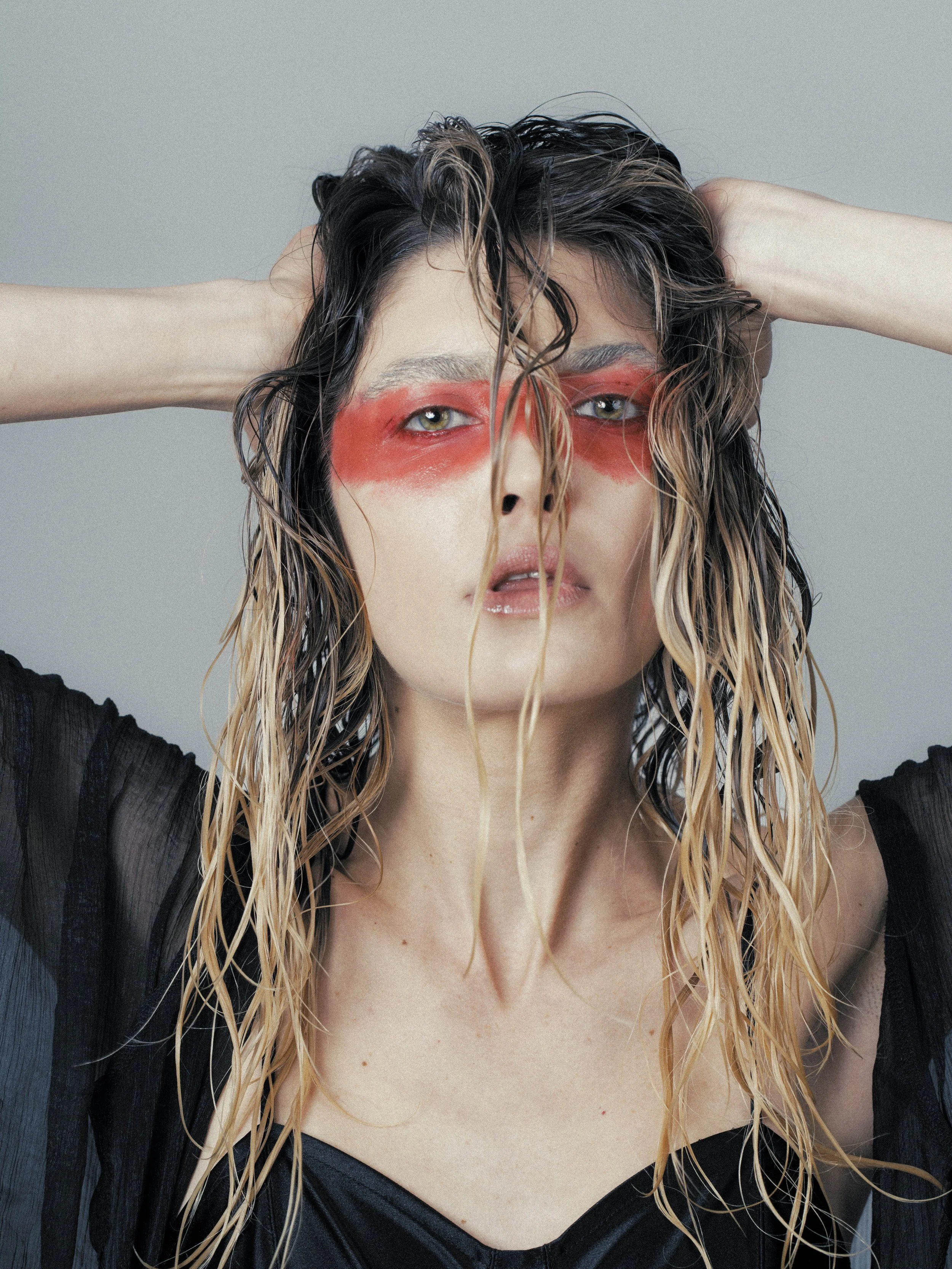A woman with wet, wavy hair and neon makeup around her eyes, holding her hair back with her hands, wearing a sheer black top with visible bra straps, against a plain gray background.
