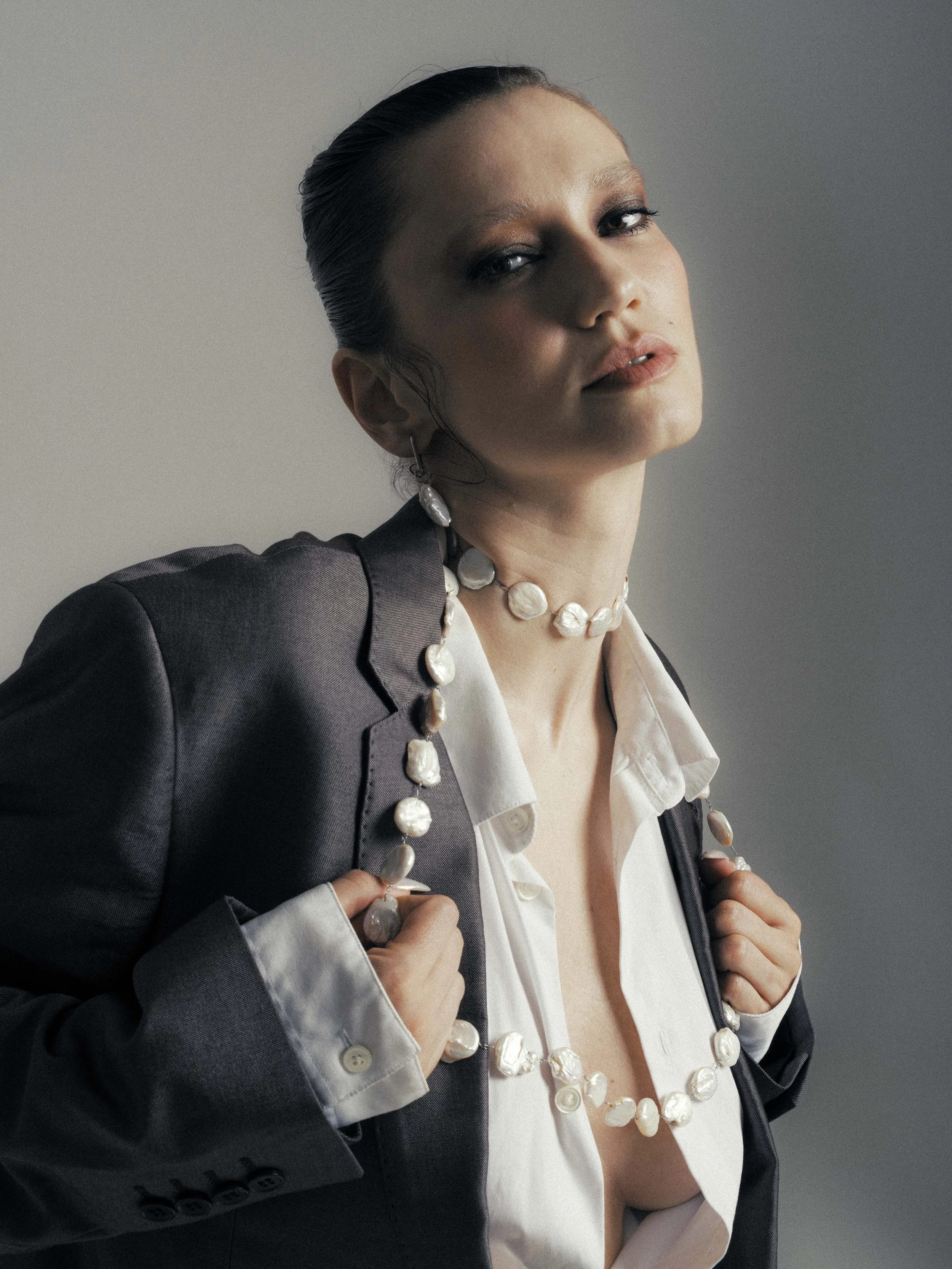 A woman wearing a dark blazer, white shirt, and a pearl necklace, with her hair slicked back, holding the lapels of her blazer, looking confidently at the camera.