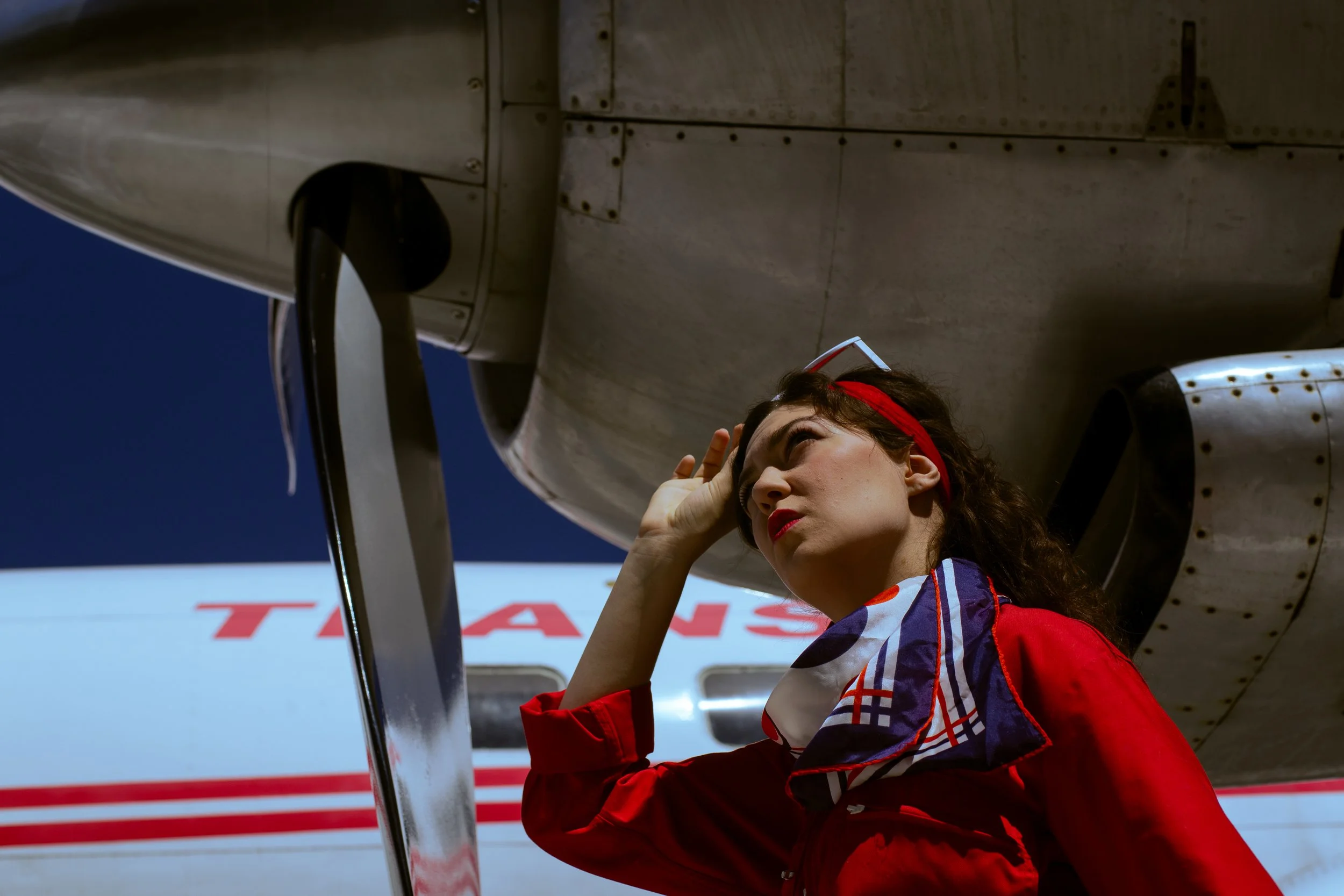 A woman with dark hair, wearing a red headband and a colorful racing jacket, is adjusting her goggles beneath the wing of an airplane at an airport or racing event.