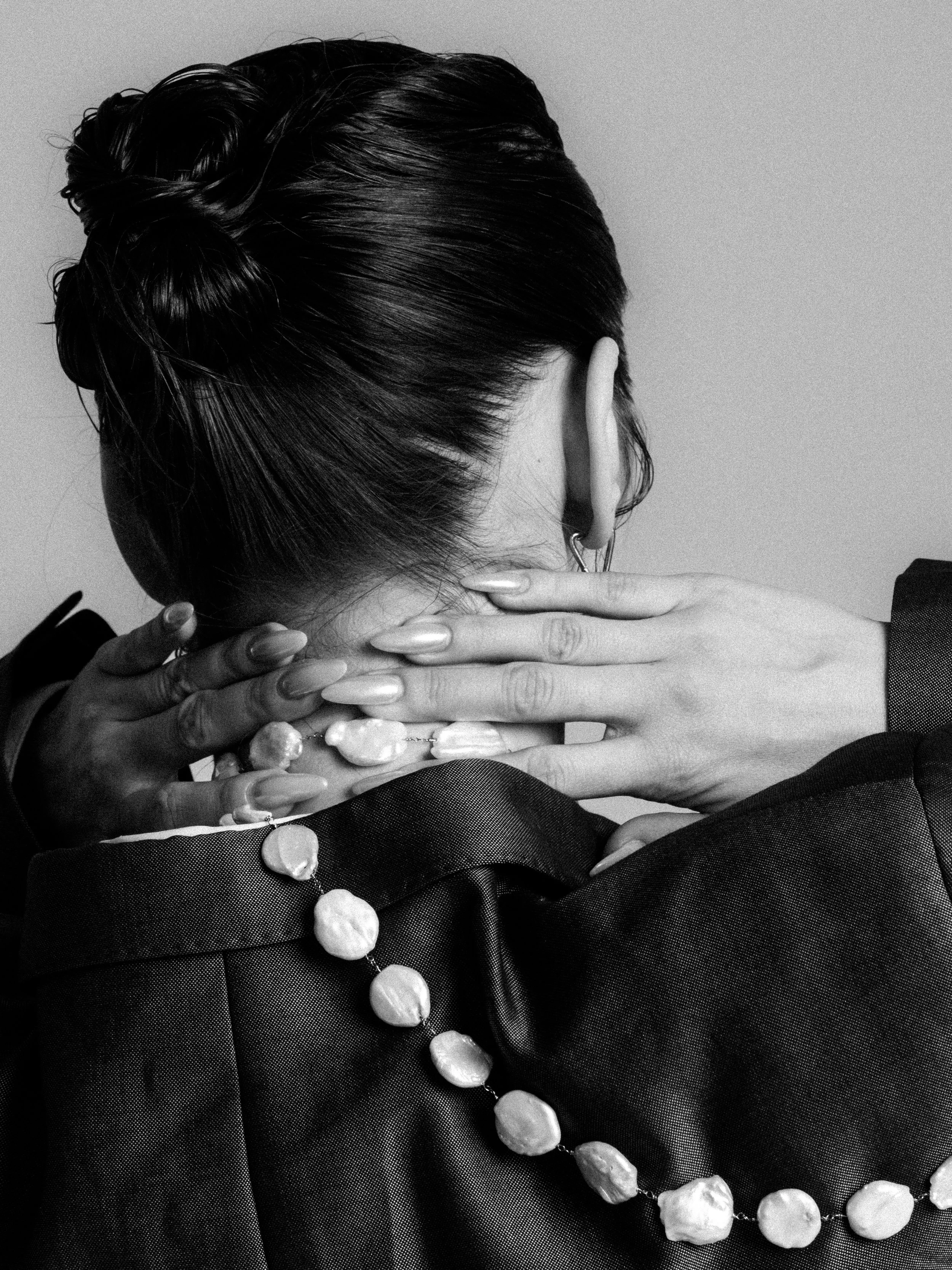 A black and white photo of a woman with her head turned away, showing her hair styled in an elegant updo. She is wearing a pearl necklace and matching pearl earrings. A person with darker skin and manicured nails is seen adjusting her necklace and ha
