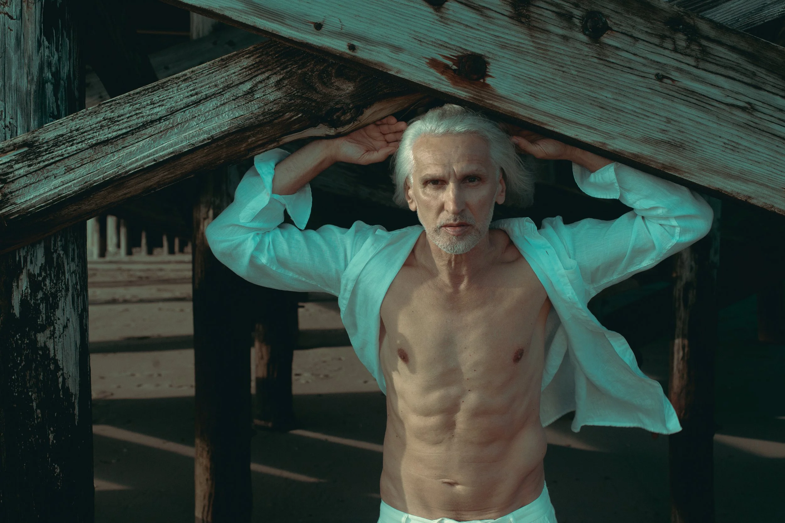 An older man with long white hair, shirt open, exposing his chest and abdomen, standing beneath a wooden structure with weathered planks, on a sandy beach.